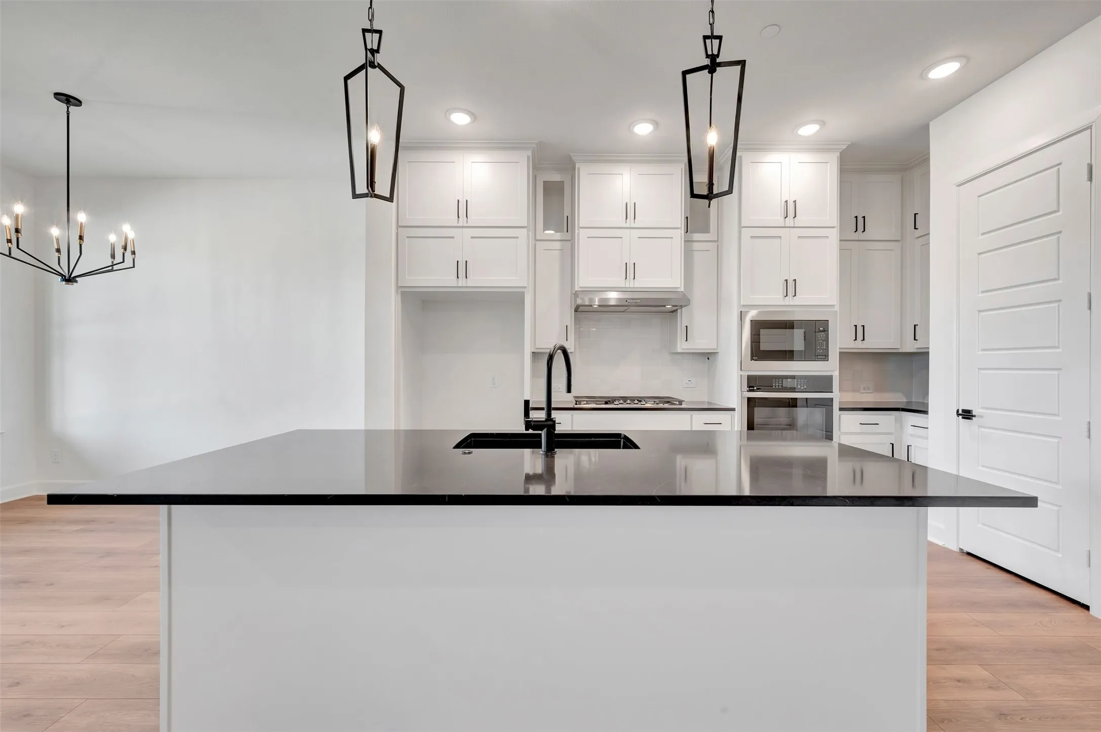 Kitchen featuring a large central island with a black countertop and integrated sink, white cabinetry with black hardware, stainless steel range hood, built-in microwave, and wood-finish flooring