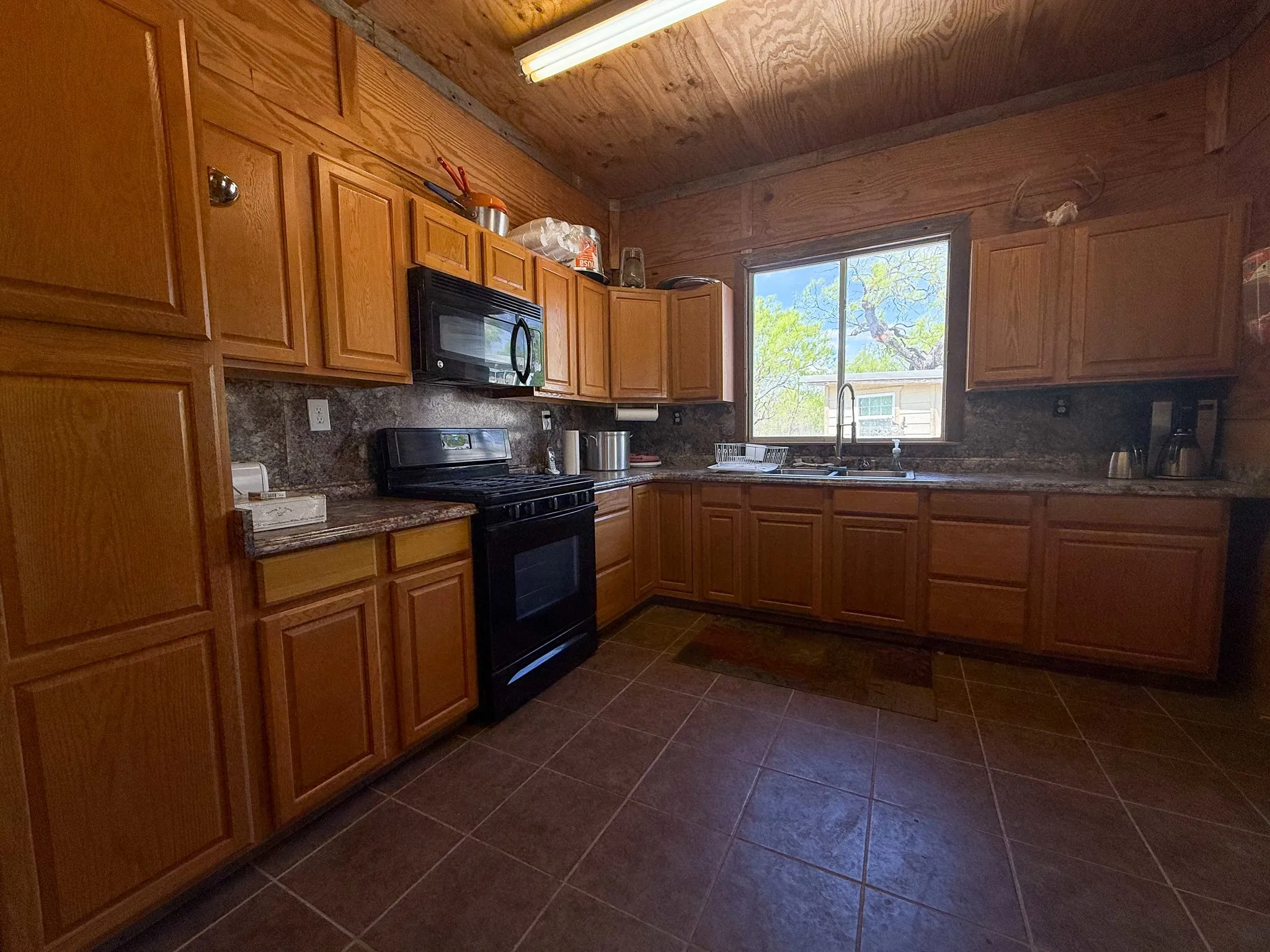 Kitchen featuring extensive wood cabinetry, L-shaped countertop space, a black range with an overhead microwave, a window above the sink, and tile flooring