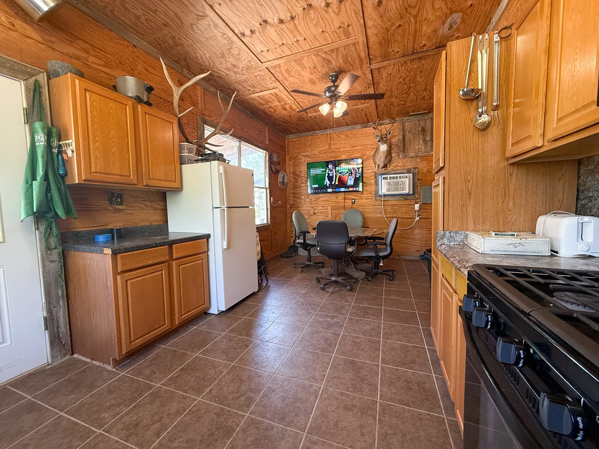 Functional kitchen area featuring wood-paneled walls and ceiling, brown square tile flooring, wood-finish cabinetry, and black countertops