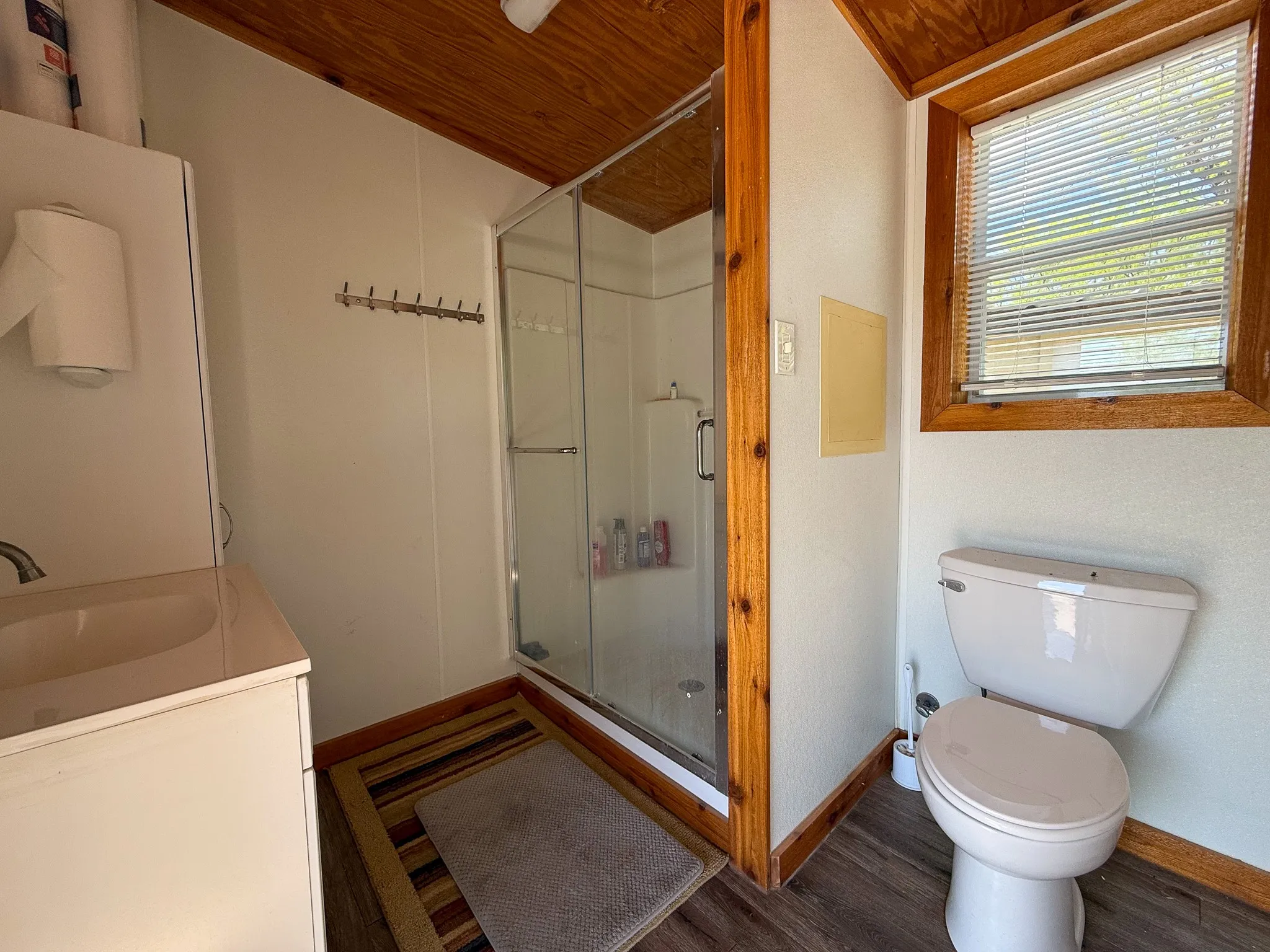 Compact bathroom featuring a glass-enclosed shower, single vanity with integrated sink, wood-finish flooring, a wood-paneled ceiling, and a window with blinds