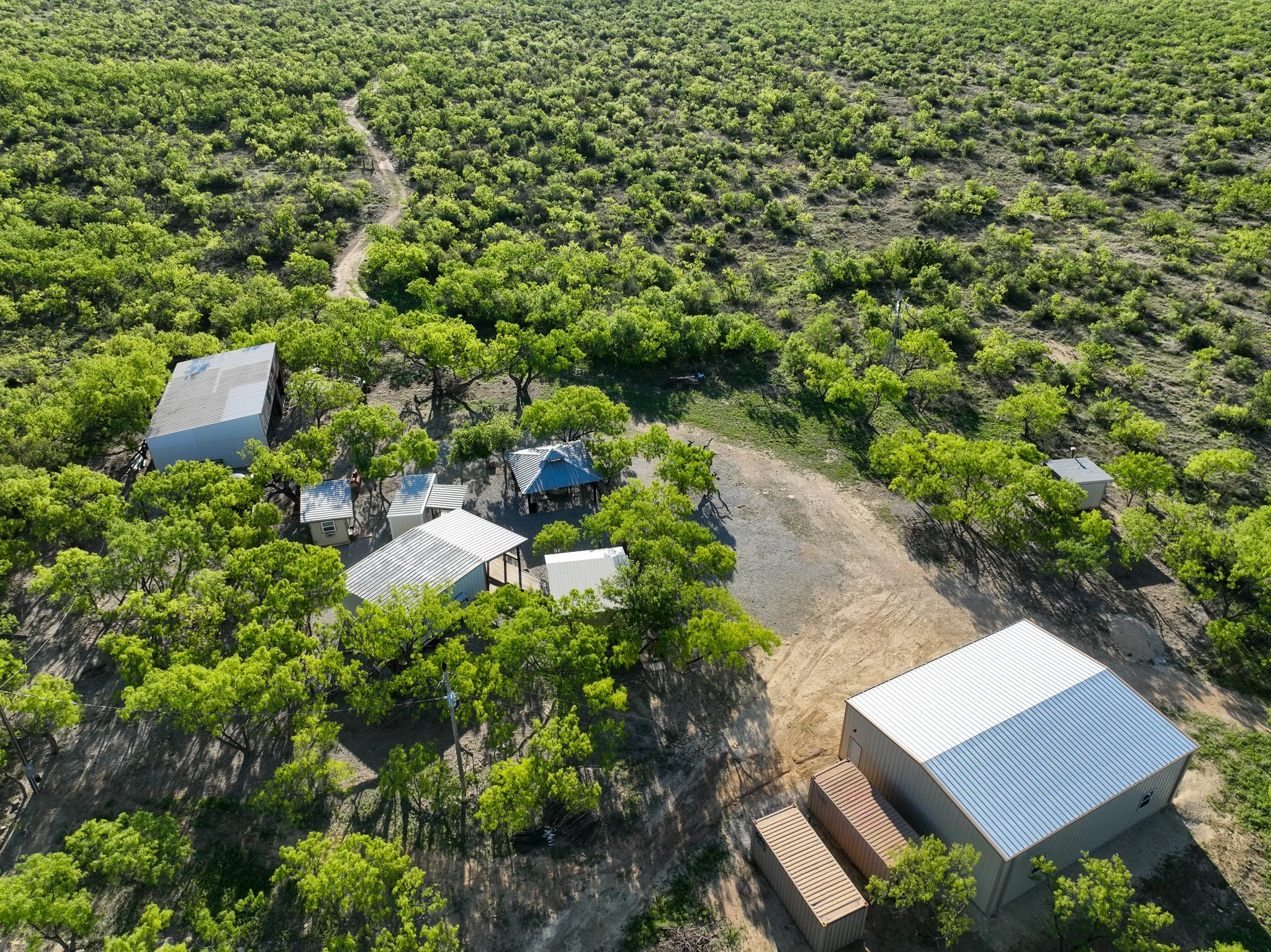 Expansive property featuring multiple metal-roofed structures, a large warehouse building, and a series of container units within a natural, tree-dotted landscape