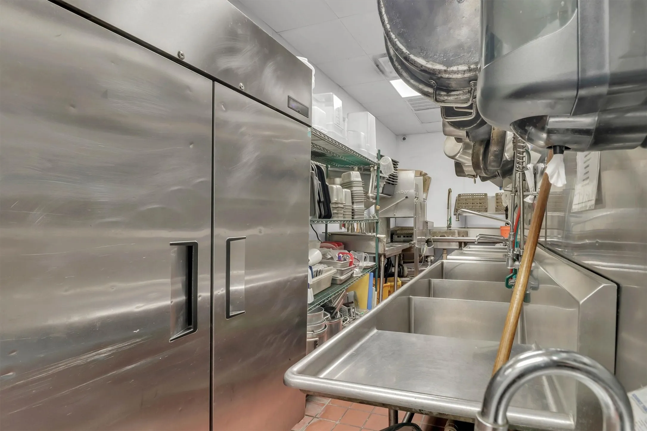 Commercial kitchen featuring a multi-basin stainless steel sink, large stainless steel refrigerator, and extensive metal shelving