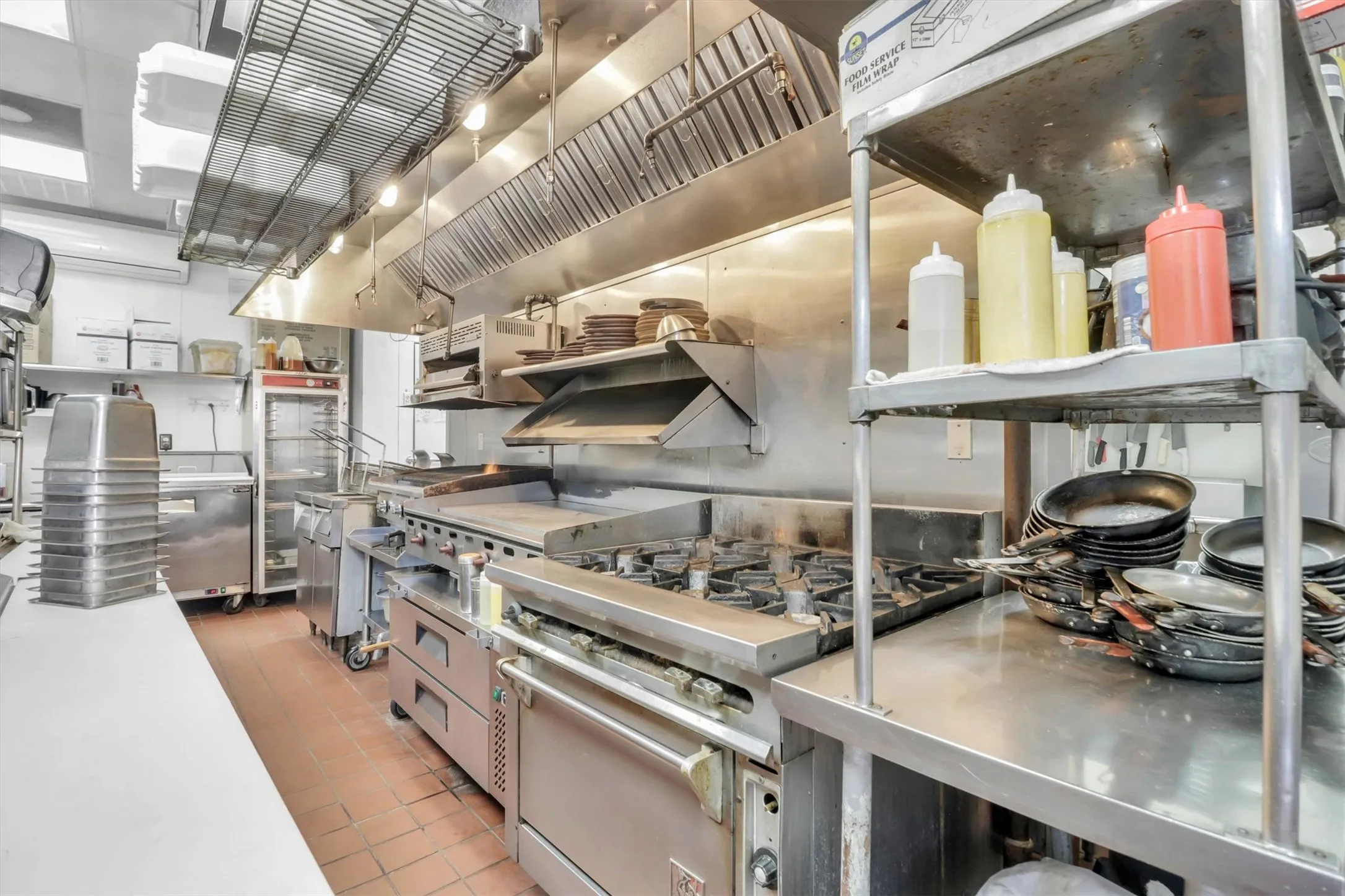 Commercial kitchen featuring a gas range with griddle, stainless steel shelving, tiled flooring, and overhead ventilation system
