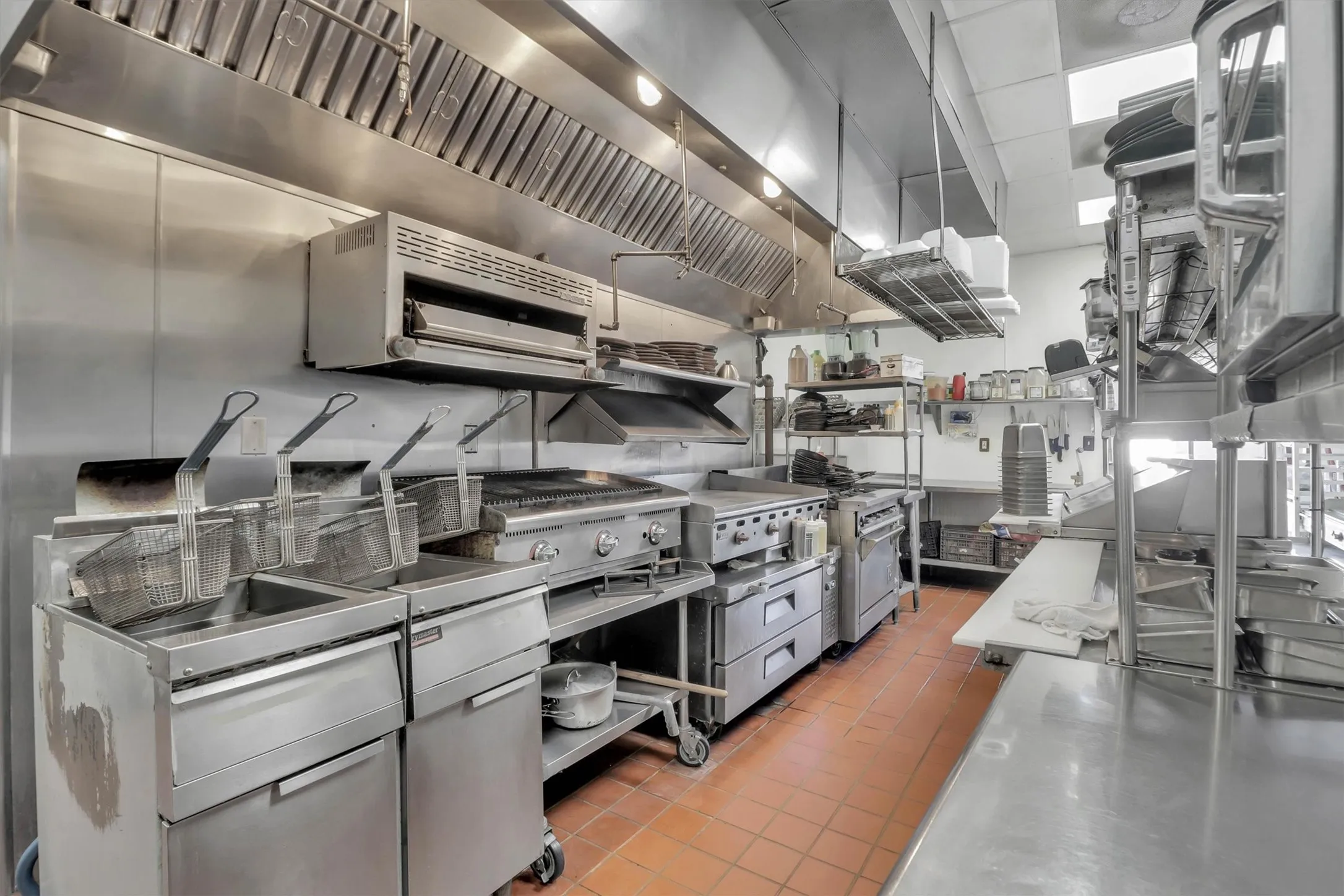 Commercial kitchen featuring stainless steel equipment, professional-grade fryers, a large range with griddle, extensive stainless steel countertops, and a full-wall exhaust hood system
