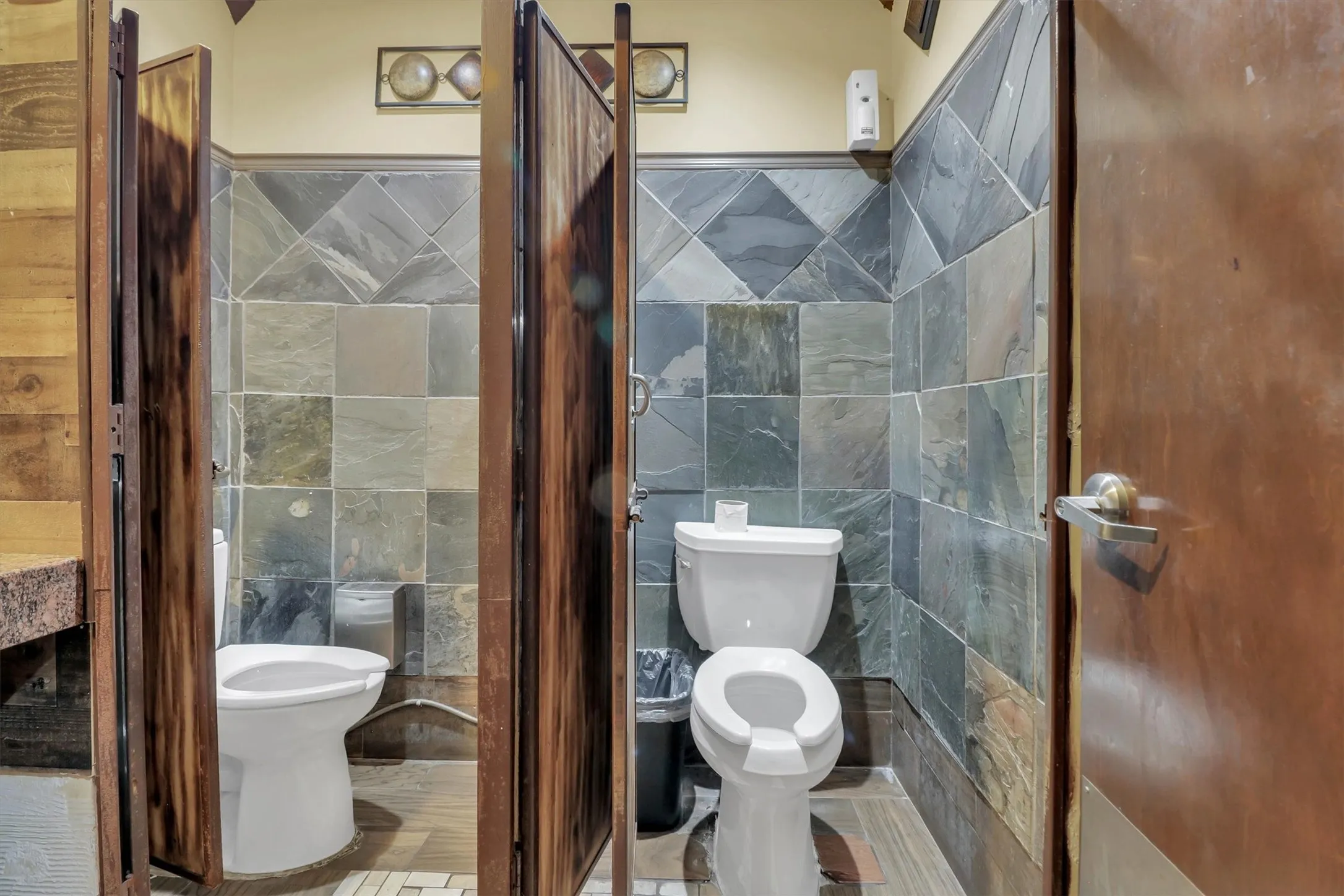 Restroom facility featuring wood-look stall doors, slate-style wall tiling, and white toilets