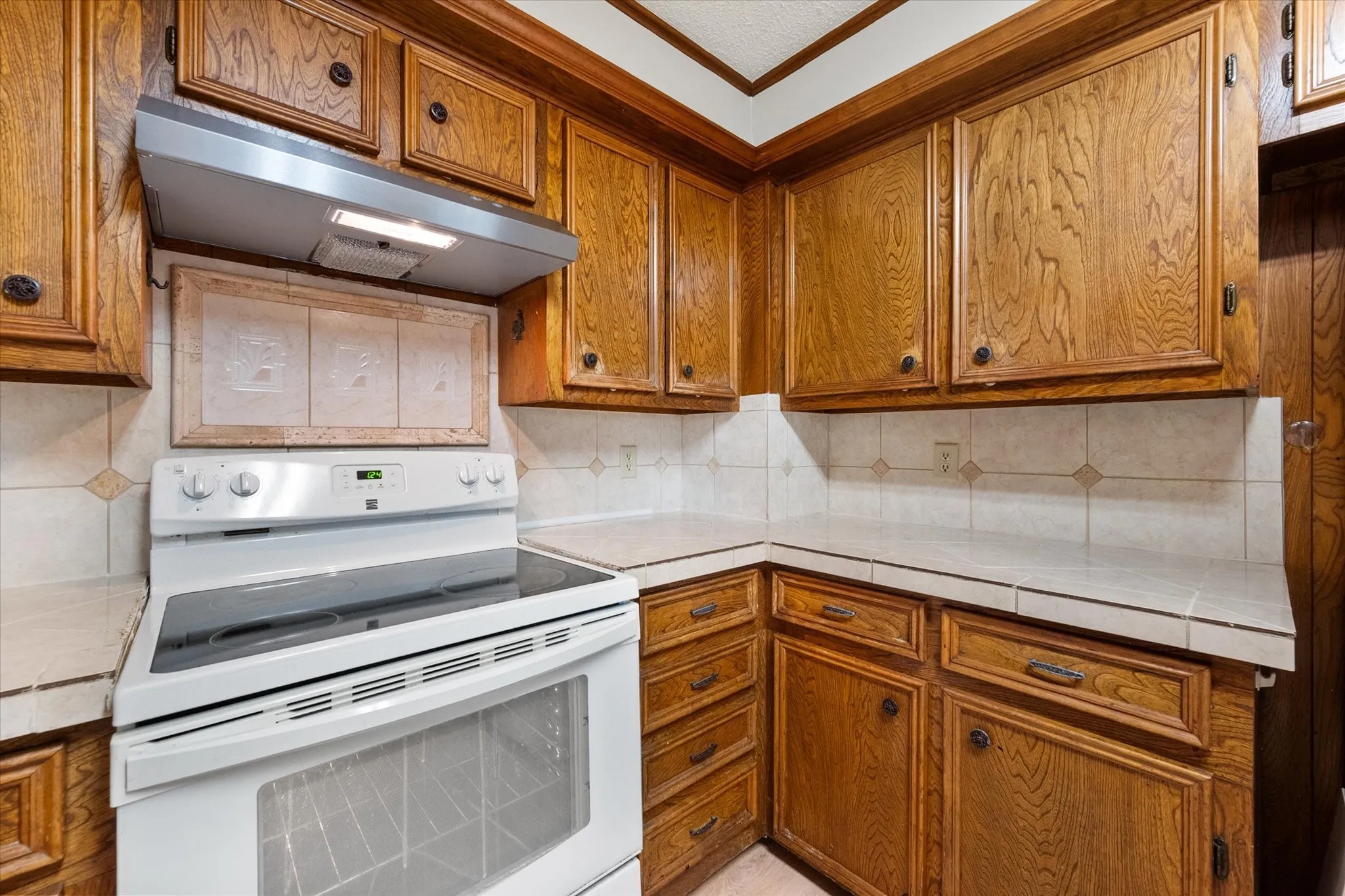 Kitchen featuring a white electric range with a stainless steel range hood, ample wood cabinetry, tile countertops, and a tile backsplash