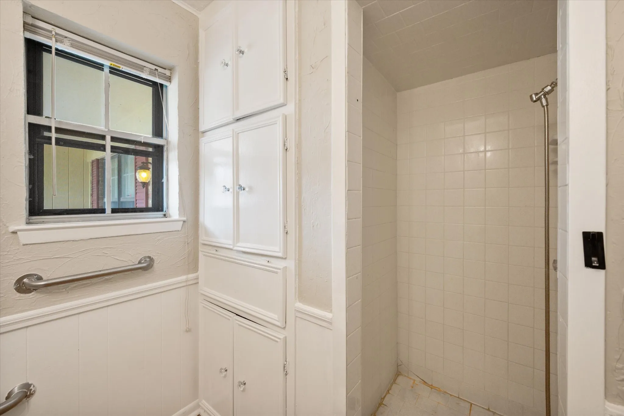 Built-in shower with white tile surround, window with black framing, integrated storage cabinetry, wainscoting, and grab bar
