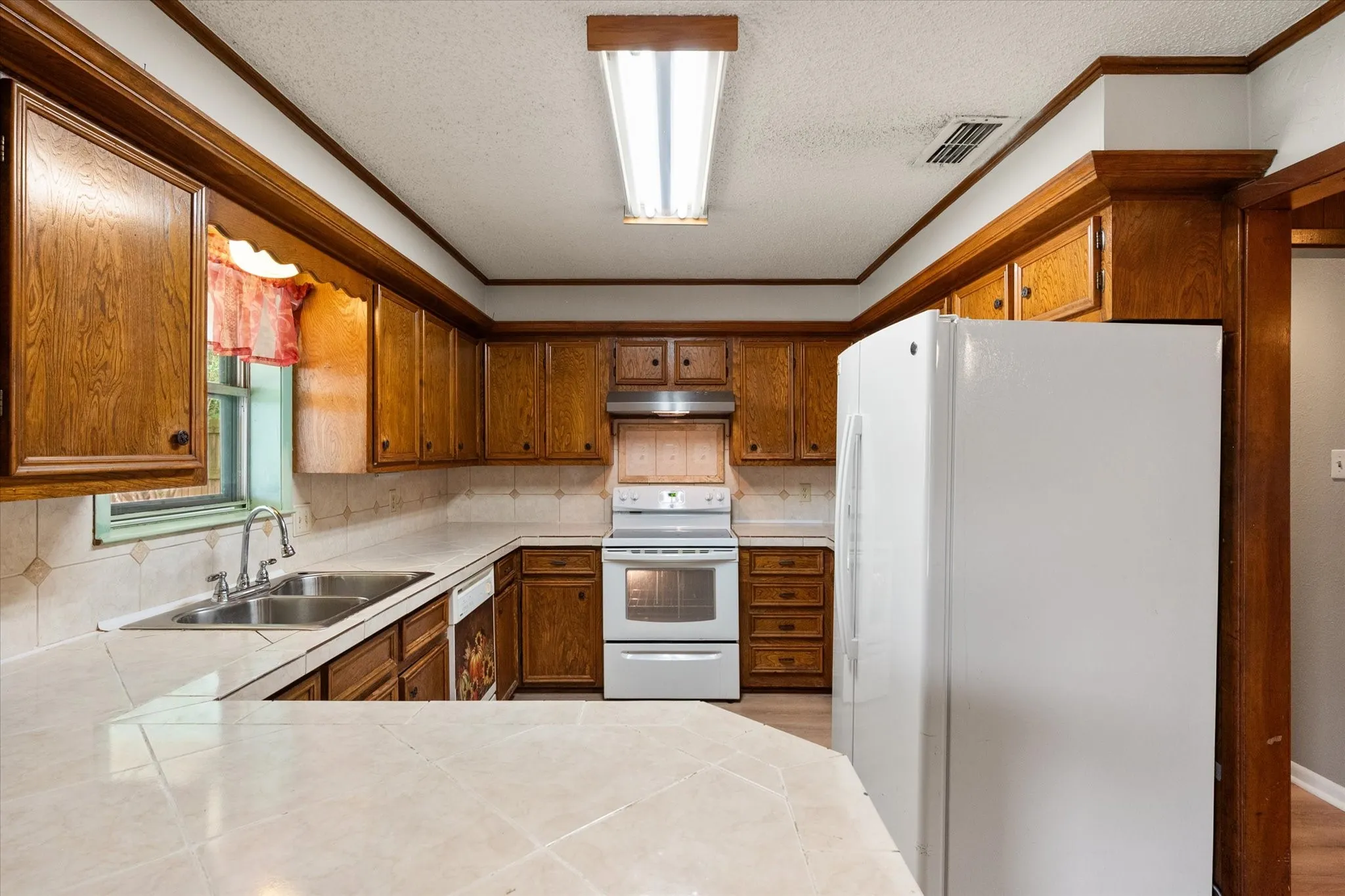 Kitchen featuring wood cabinetry, tile countertops, a double basin stainless steel sink, and white appliances including a range and refrigerator