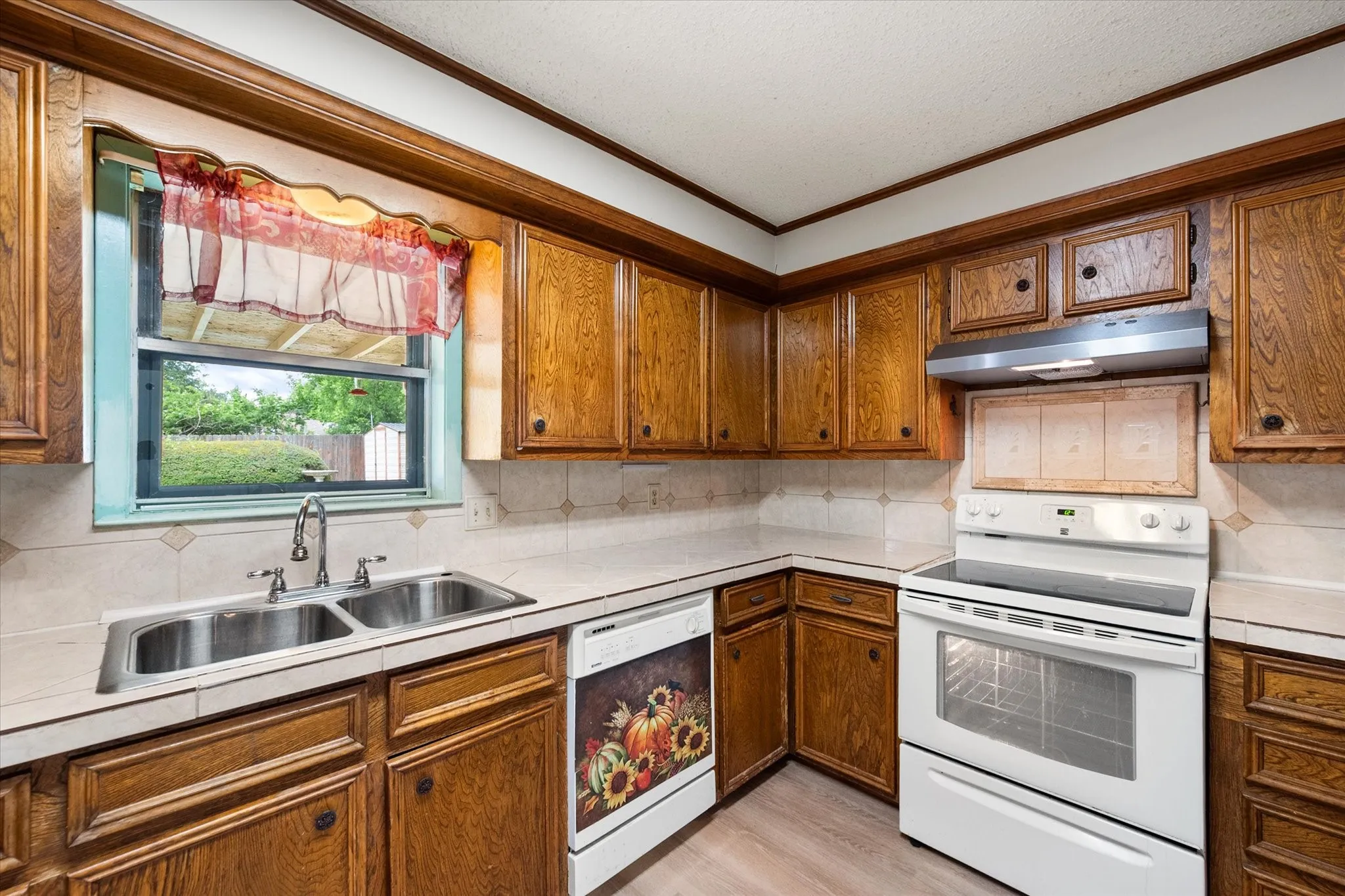 Kitchen featuring wood-finish cabinetry, tile countertops, a double basin stainless steel sink, and a white electric range with an overhead vent hood