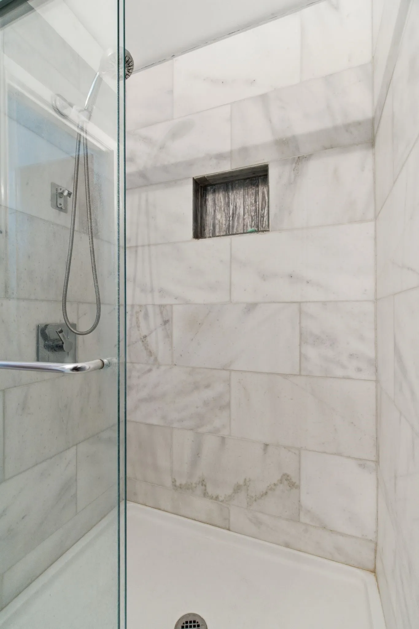 Glass-enclosed shower featuring white tiled walls, a built-in shower niche, and a white shower pan with a chrome drain