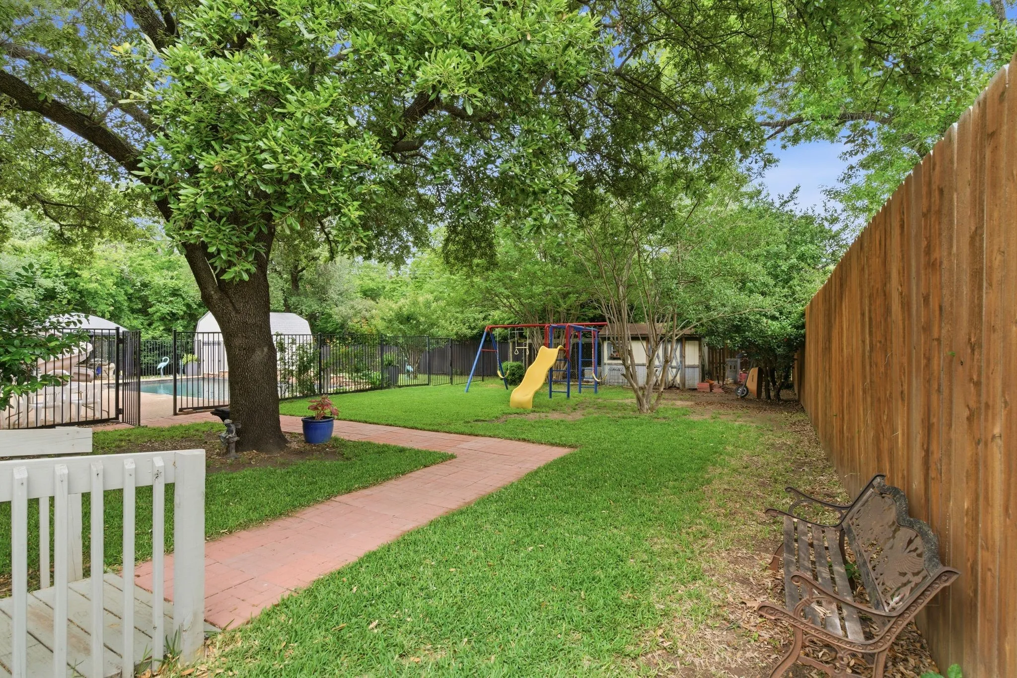 Expansive backyard featuring a red brick pathway, mature trees, a wood privacy fence, and a fenced swimming pool area