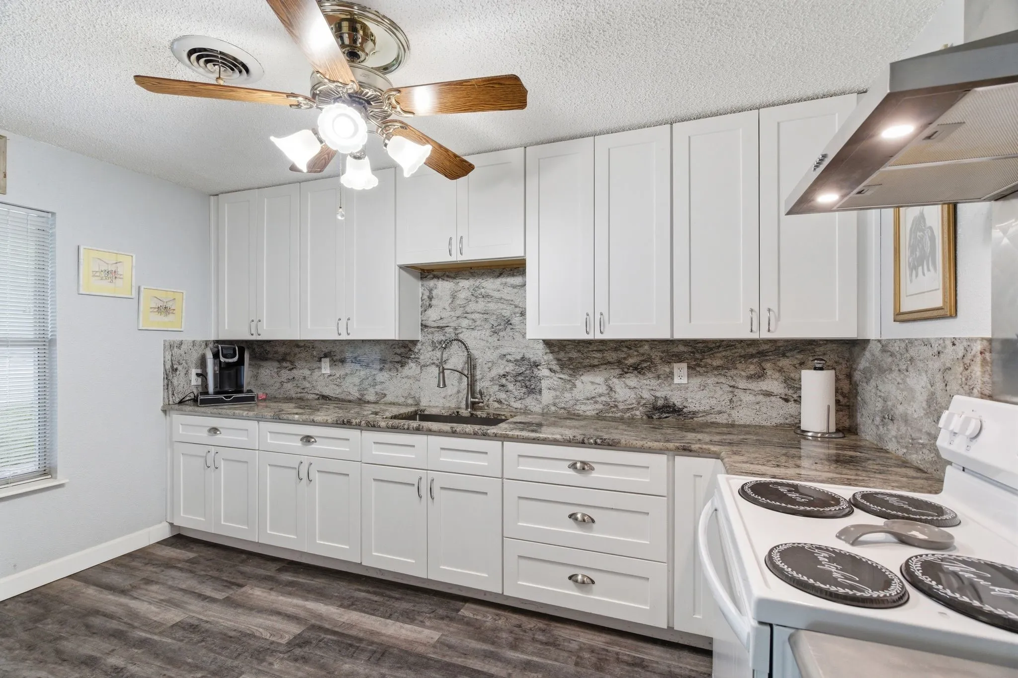 Contemporary kitchen featuring white shaker cabinetry, patterned stone countertops and backsplash, wood-finish flooring, a stainless steel faucet, and a ceiling fan with light fixture