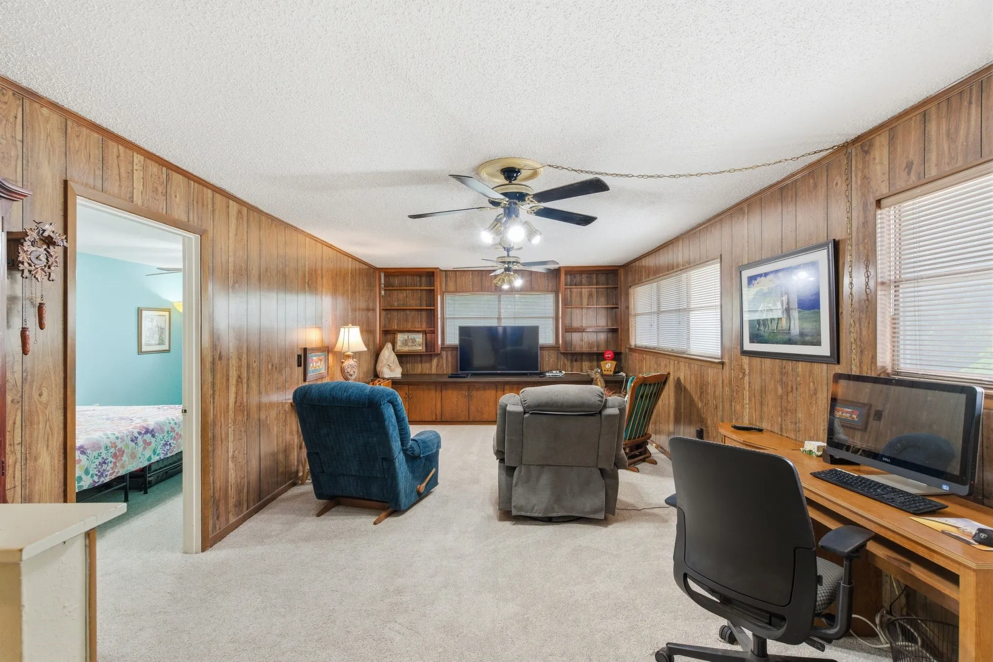 Carpeted interior space featuring wood paneling, multiple windows with blinds, a built-in media center with shelving, and ceiling fans