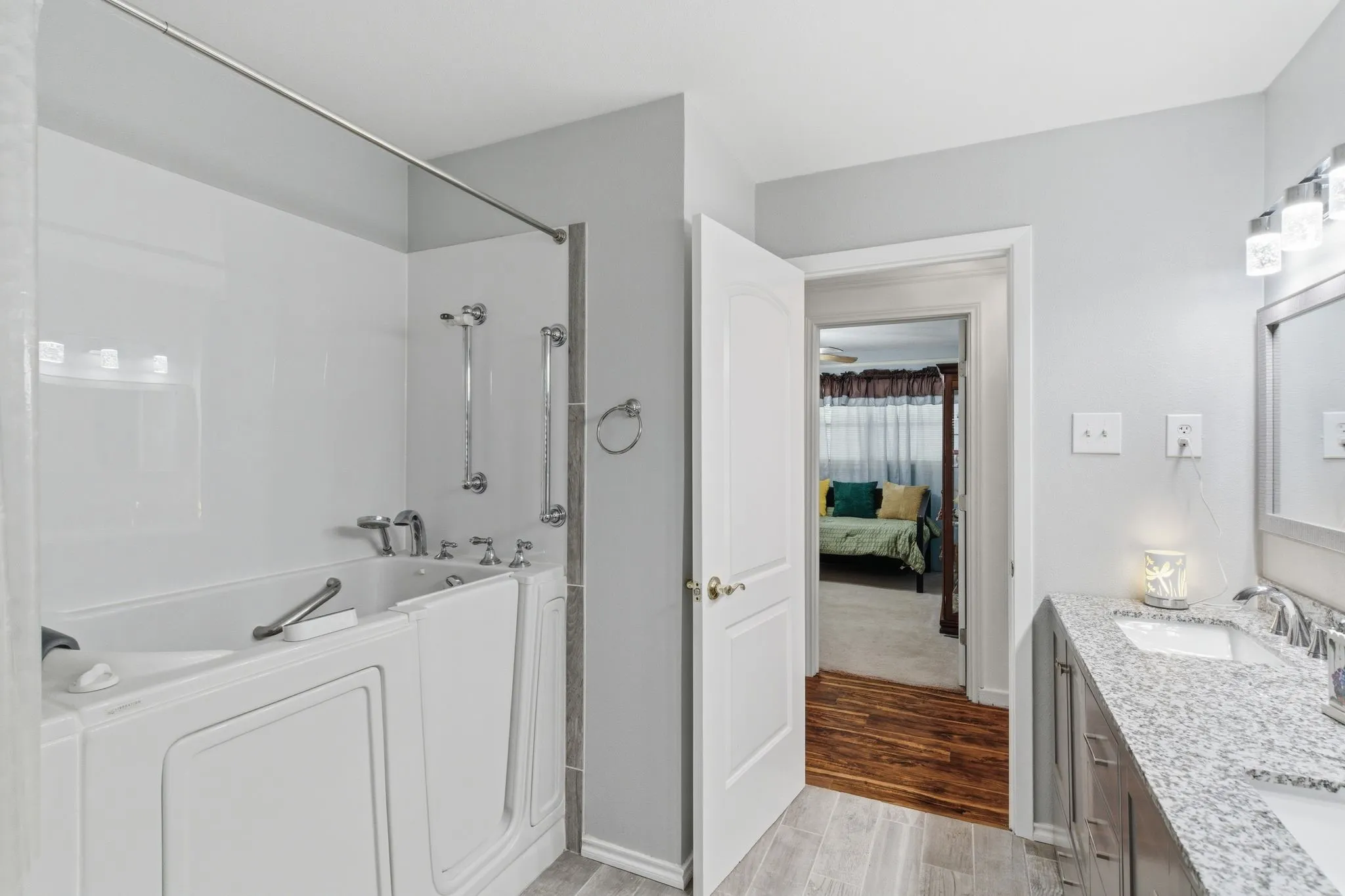 Bathroom featuring a walk-in tub with grab bars, a single-sink vanity with a granite-style countertop, and wood-finish flooring in the doorway