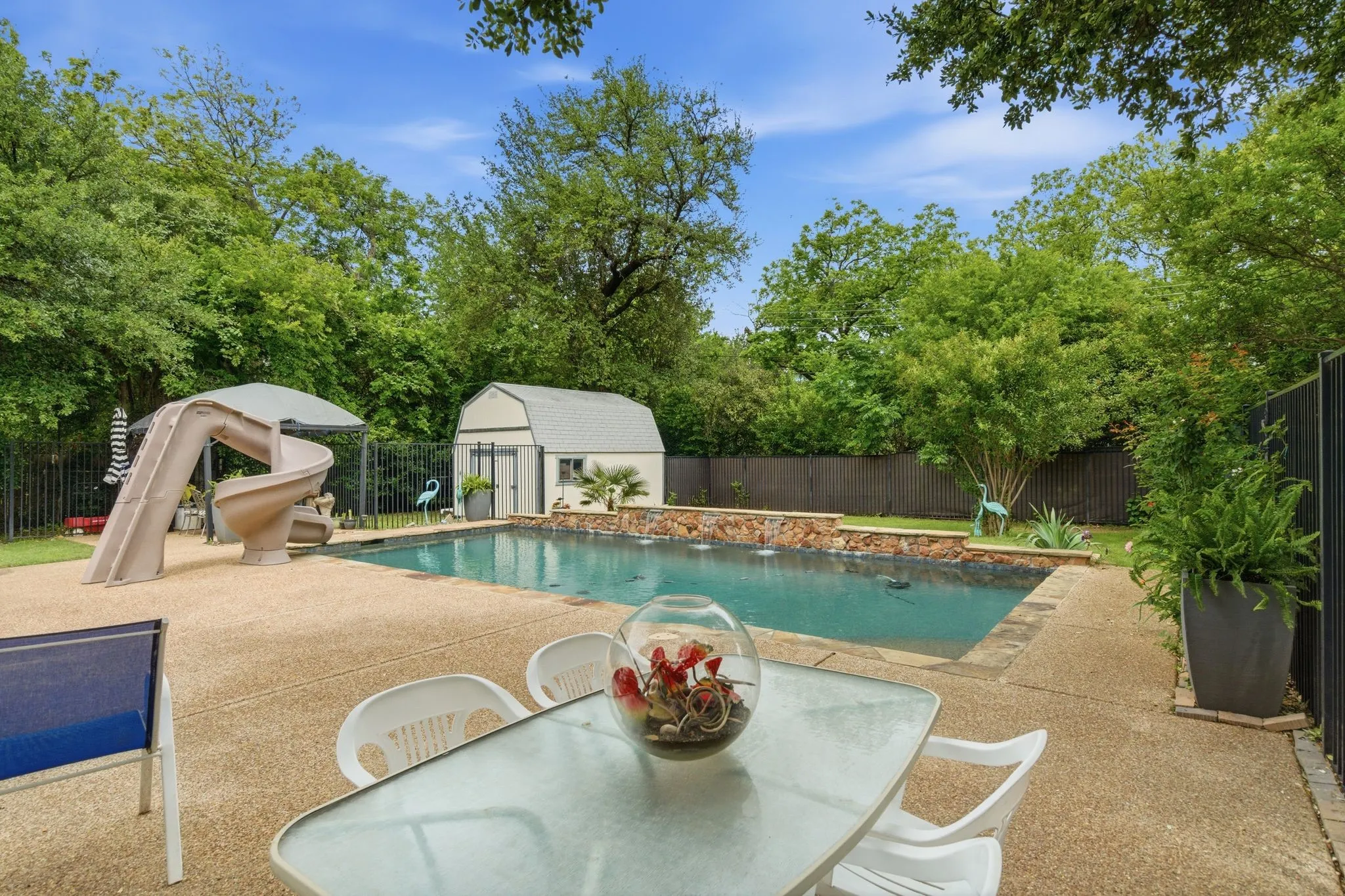 Spacious backyard featuring a rectangular swimming pool with integrated waterfall feature, large aggregate concrete patio, spiral water slide, storage shed, and mature perimeter trees