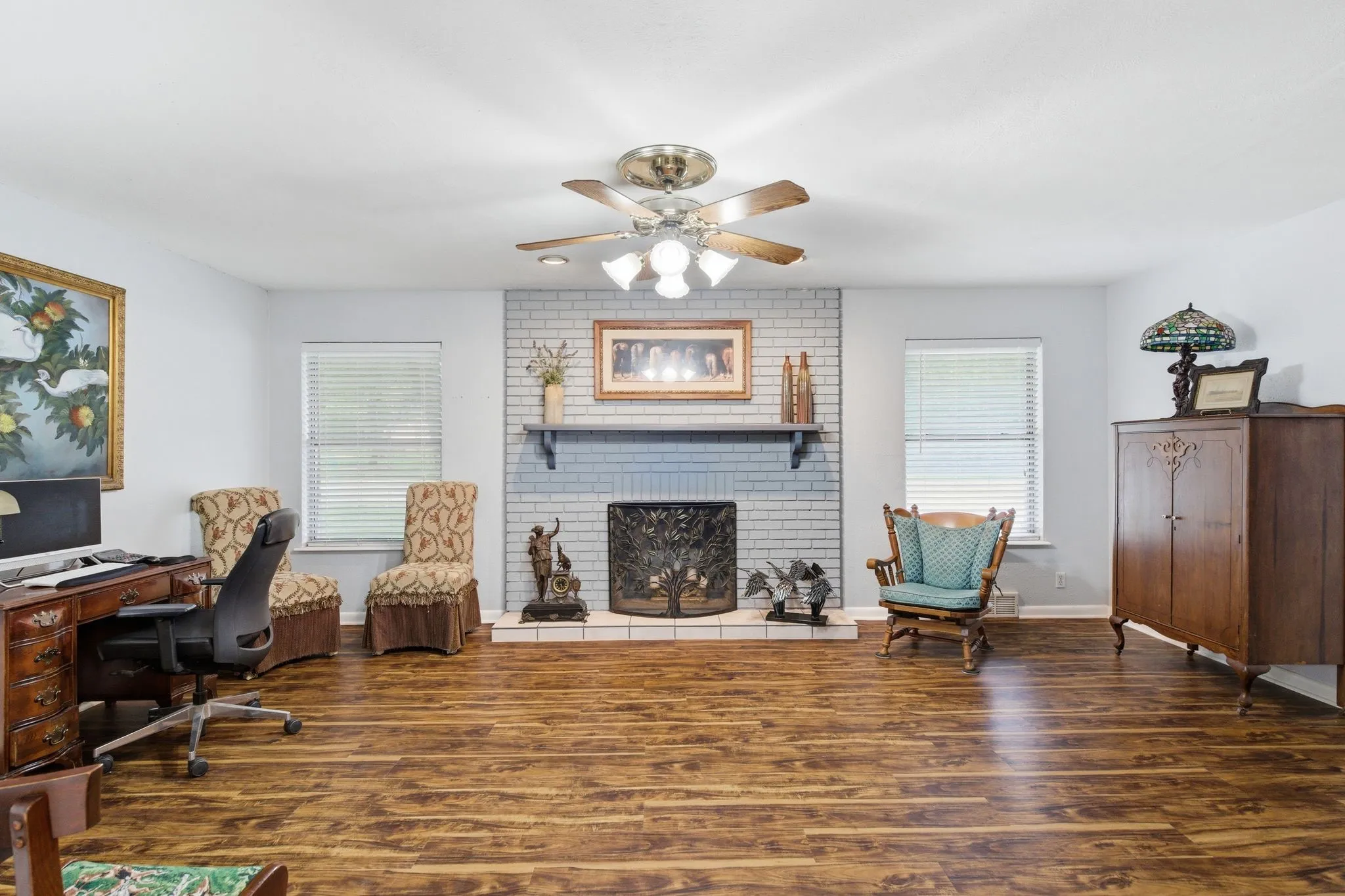 Spacious living area featuring wood-finish flooring, a painted brick fireplace, and a ceiling fan with light fixture