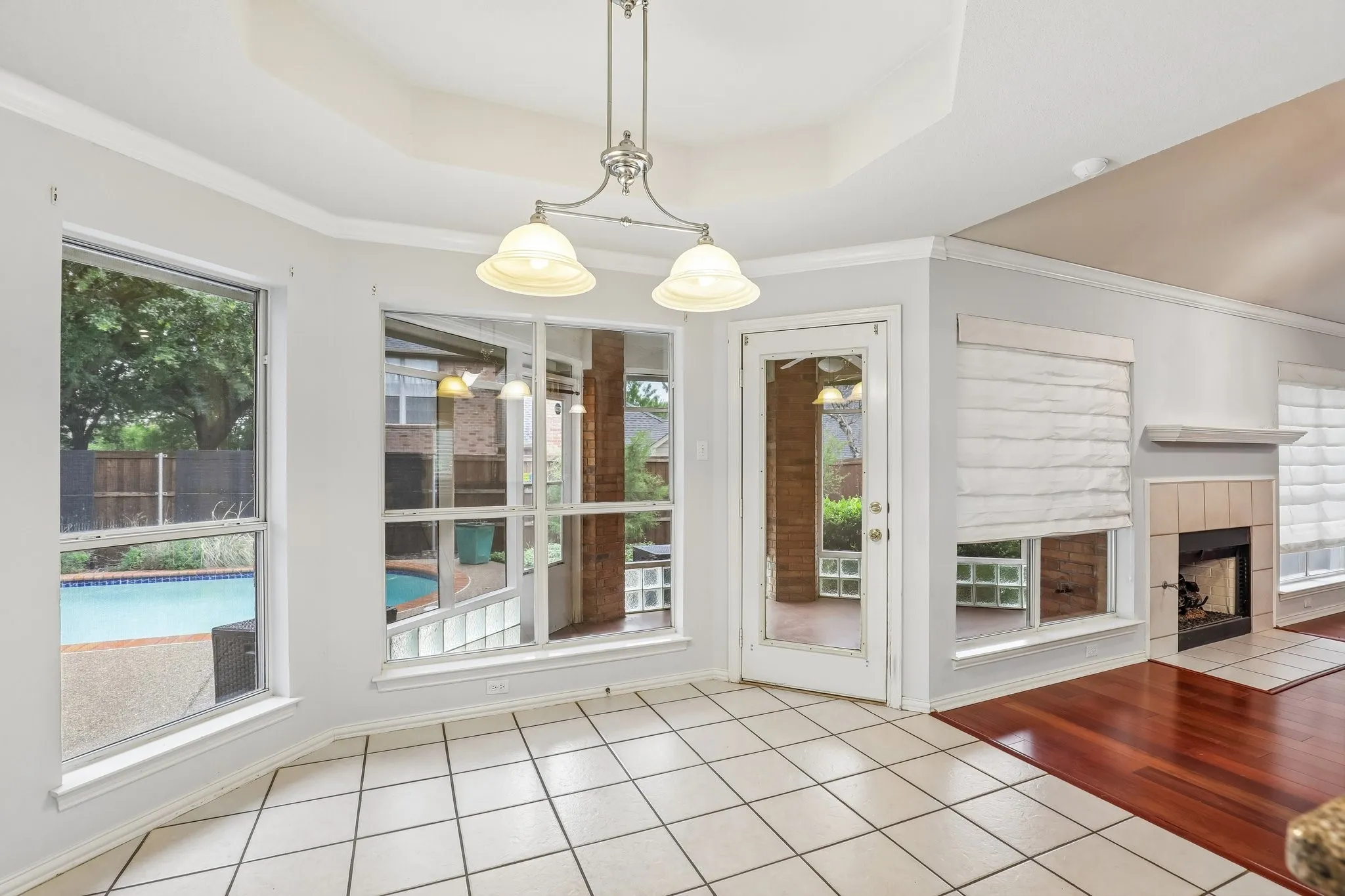 Tiled flooring in a bright dining area with an overhead chandelier, featuring multiple windows overlooking a backyard pool, a glass-paneled door, and a fireplace with a tiled hearth