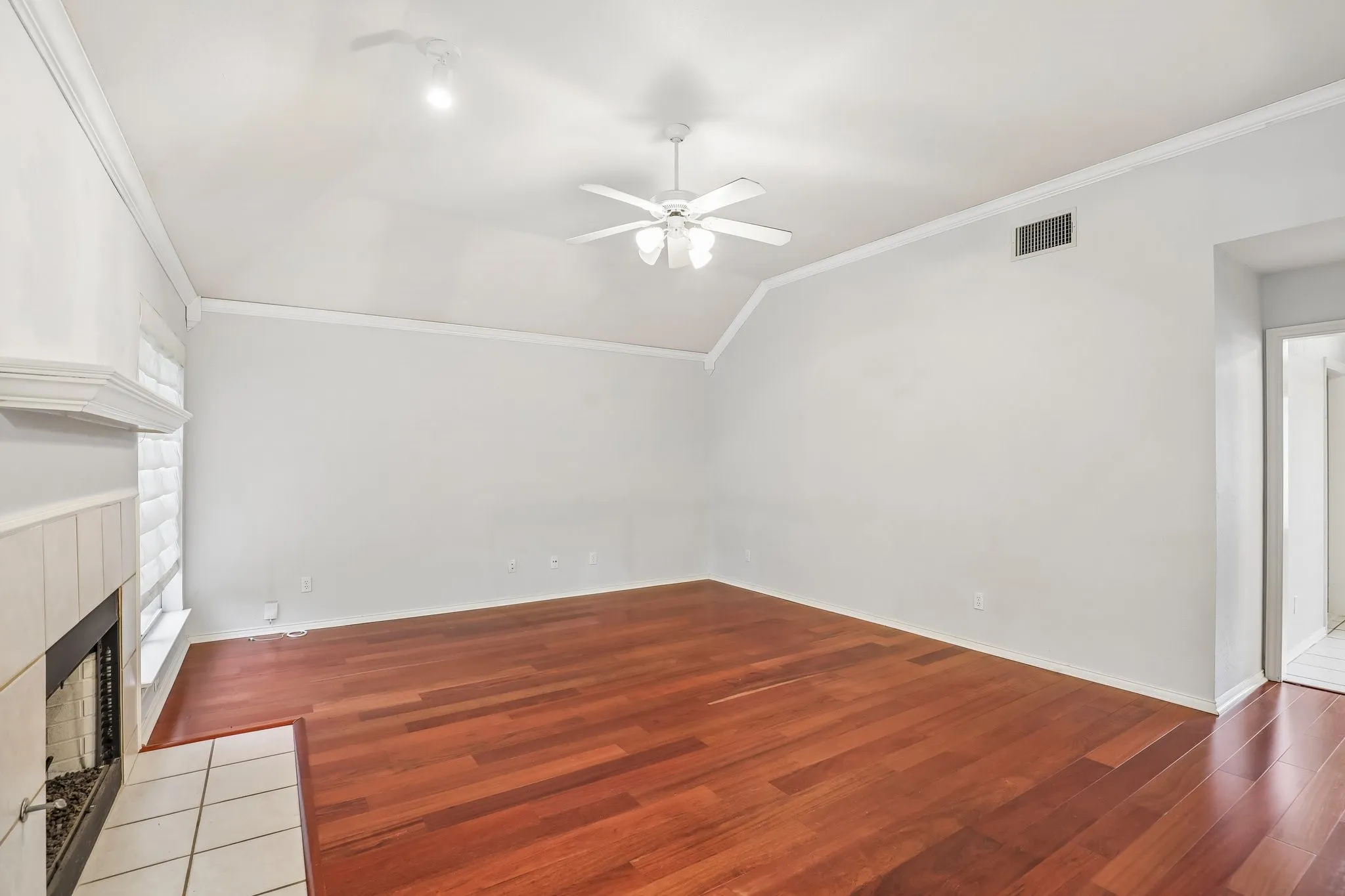 Living area featuring rich wood-finish flooring, a white tile-surround fireplace with a mantel, a vaulted ceiling, and a ceiling fan
