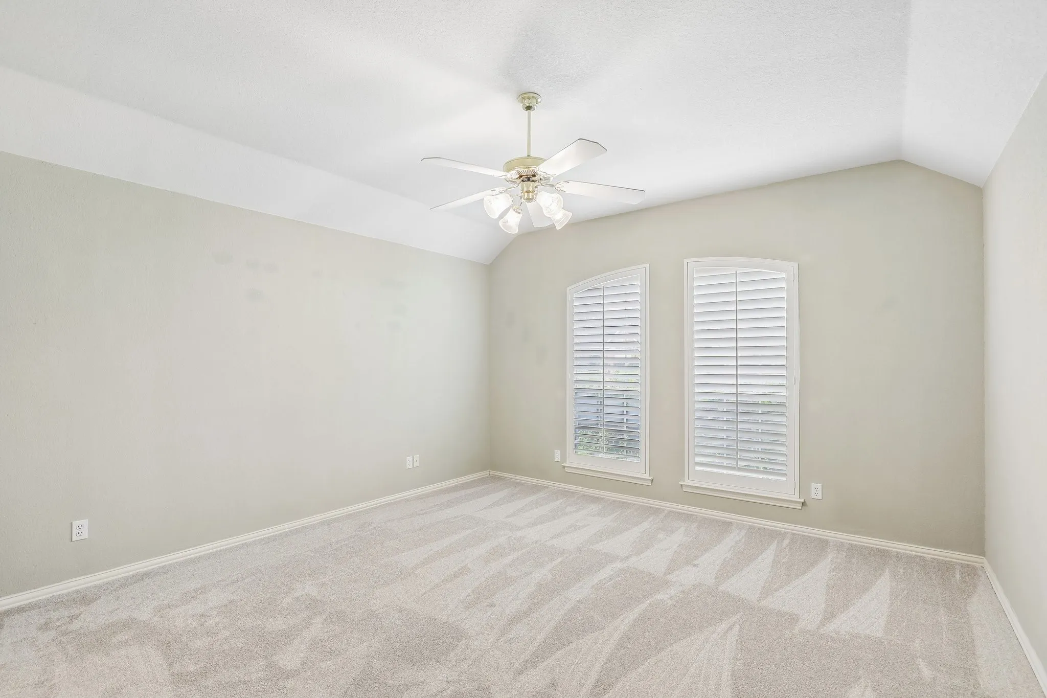 Spacious room featuring vaulted ceilings, a ceiling fan, two arched windows with louvered shutters, and light-toned carpet flooring