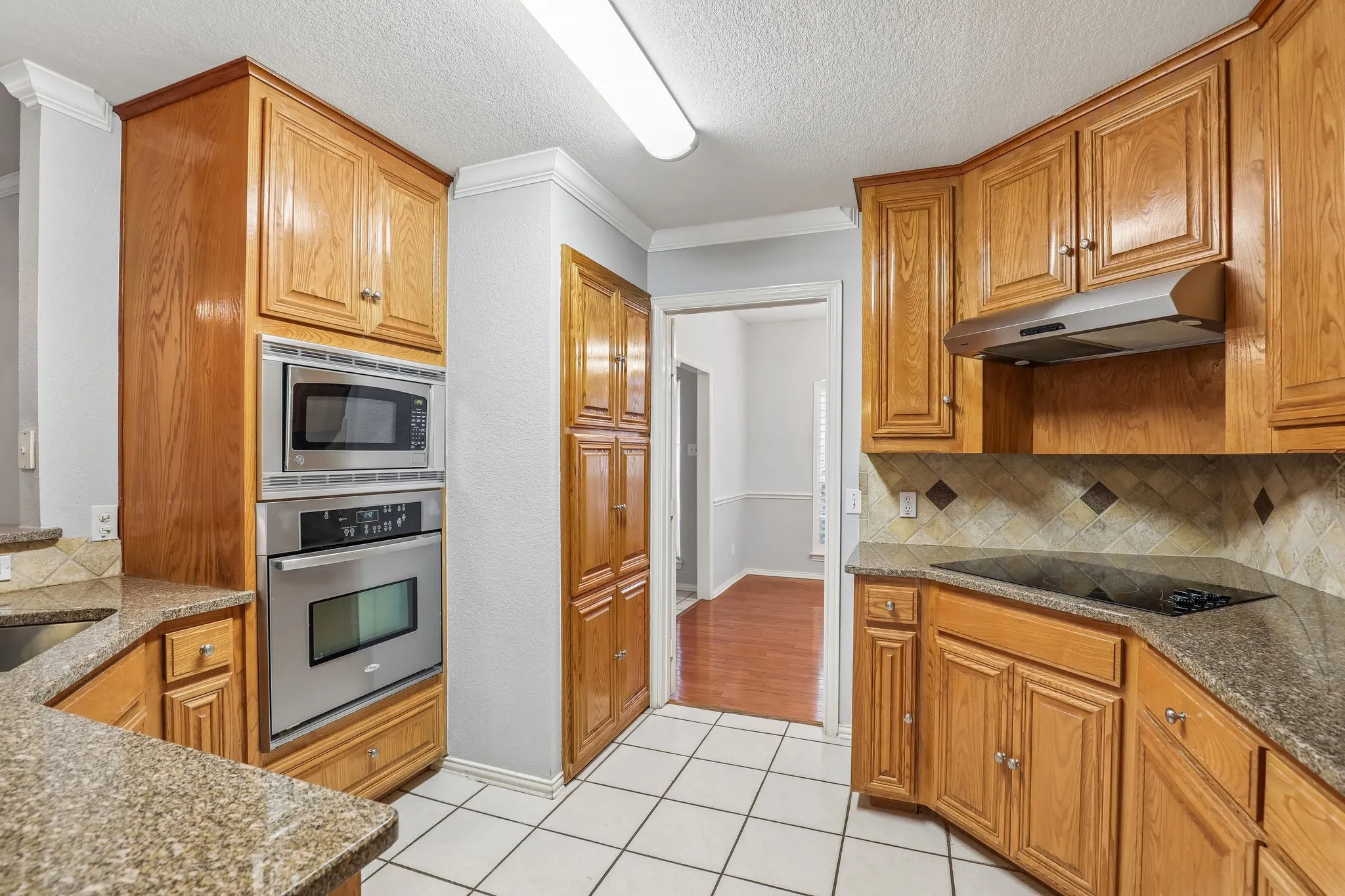 Kitchen featuring wood cabinetry, stainless steel built-in microwave and oven, a black cooktop, tiled backsplash with decorative accents, and speckled countertops
