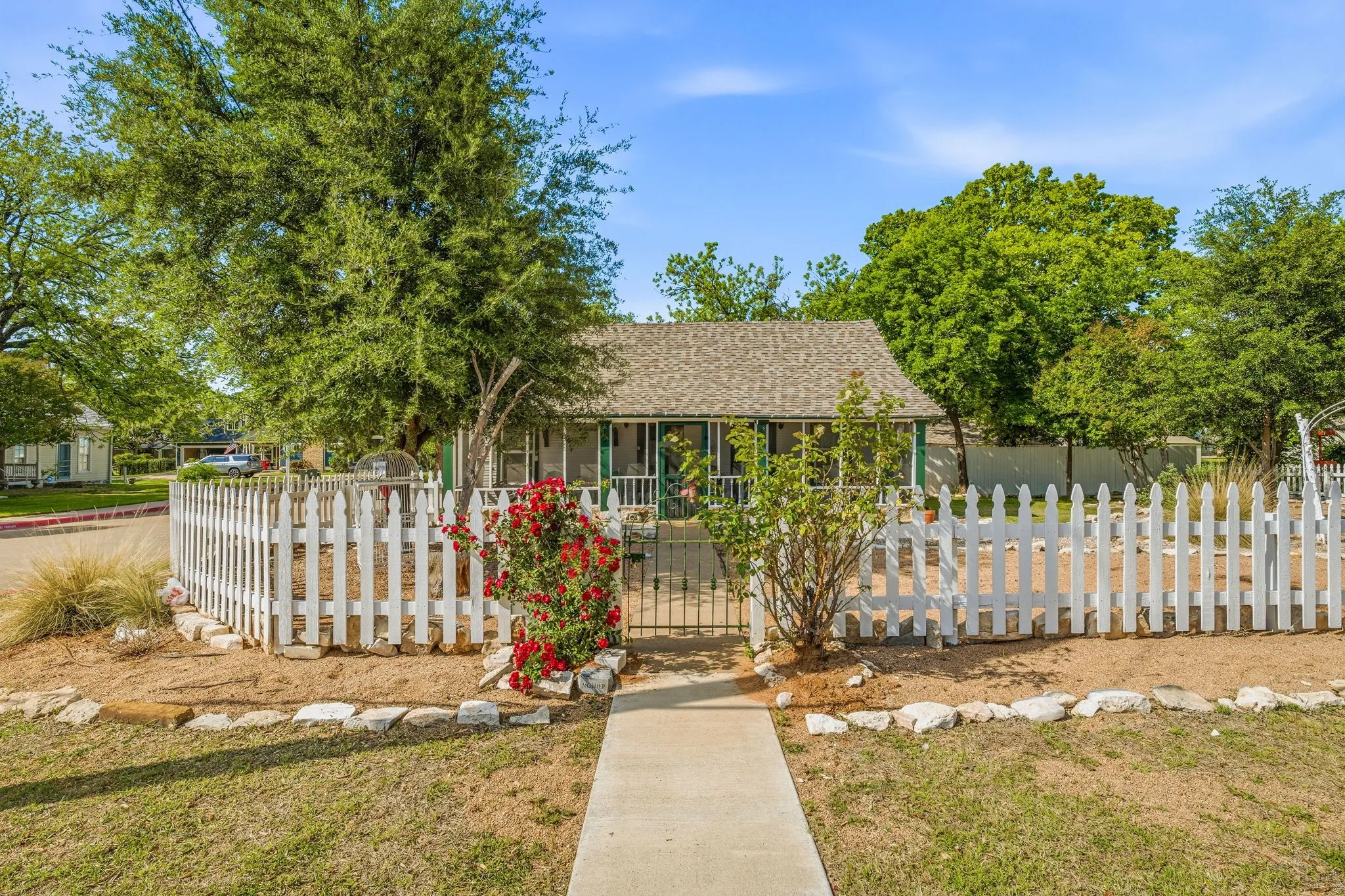 Cottage-style residence featuring a white picket fence, mature trees, a covered porch, and a concrete walkway