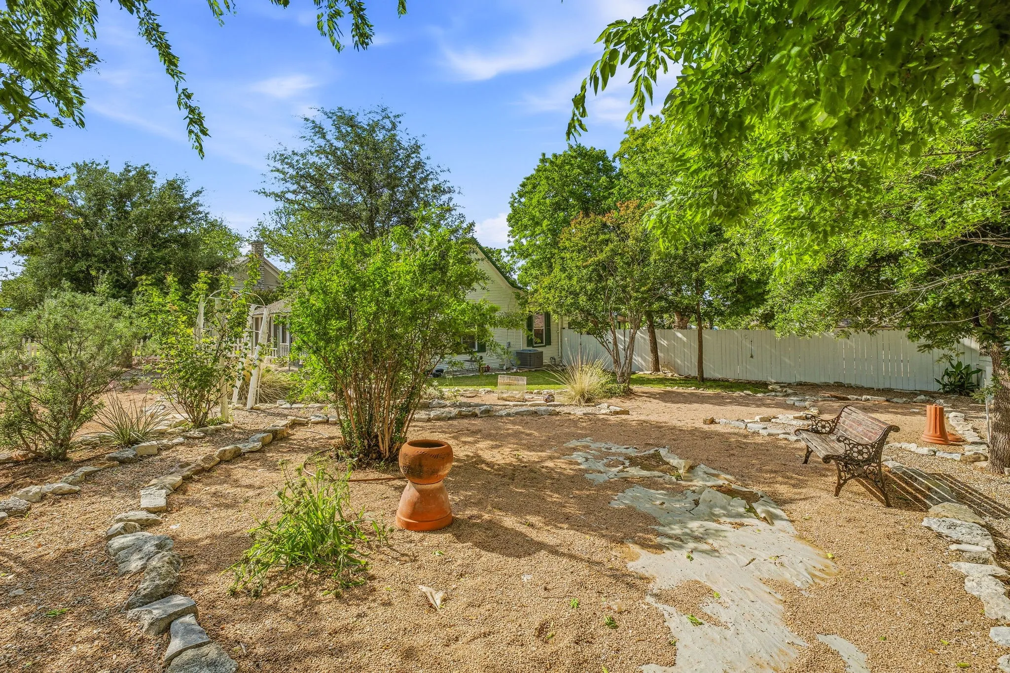 Landscaped garden area featuring gravel pathways outlined with natural stones, mature trees, various shrubs, and a decorative wrought iron bench