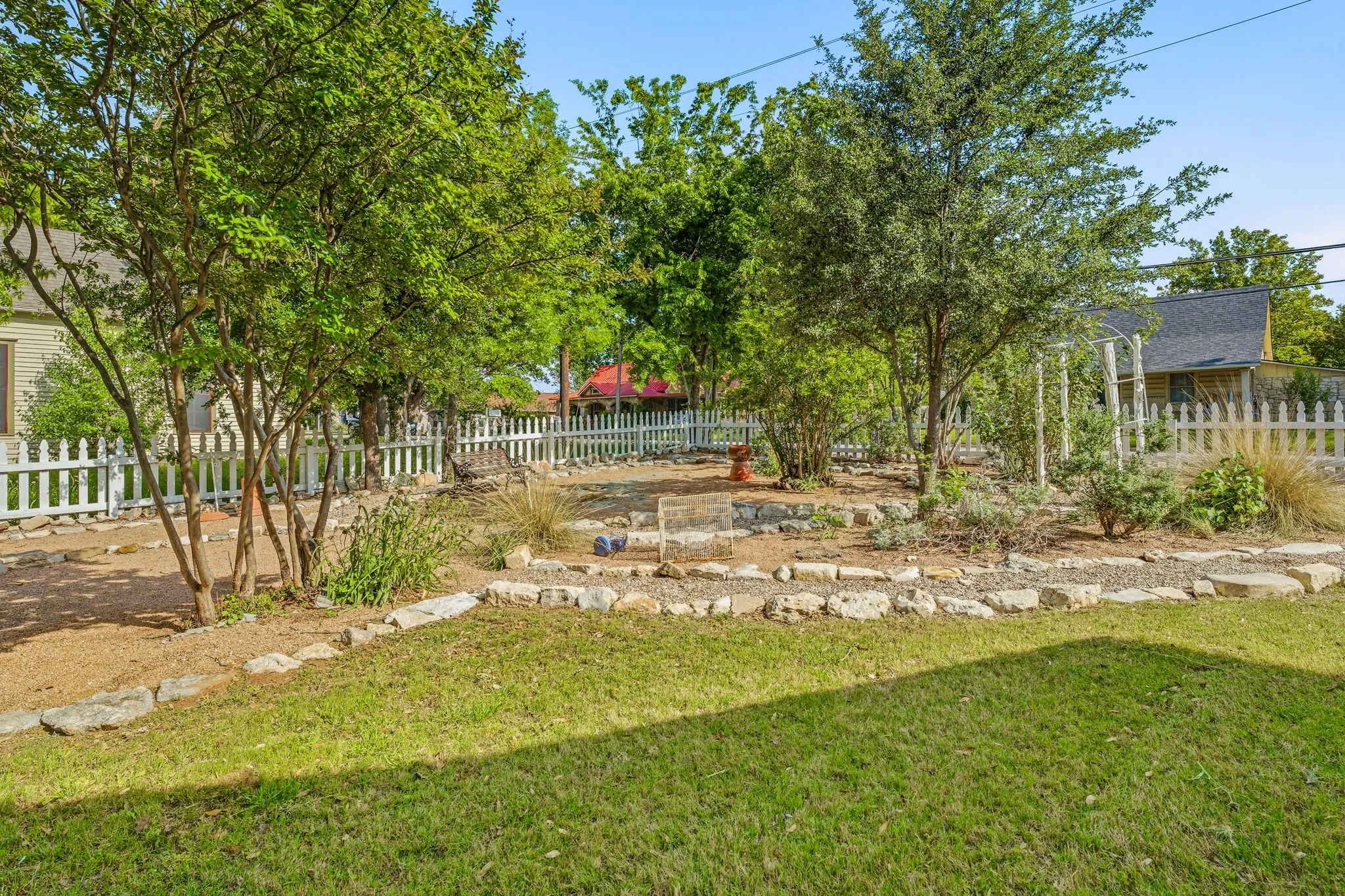 Landscaped backyard featuring a grassy area, stone-edged garden beds, numerous mature trees, and a white picket fence