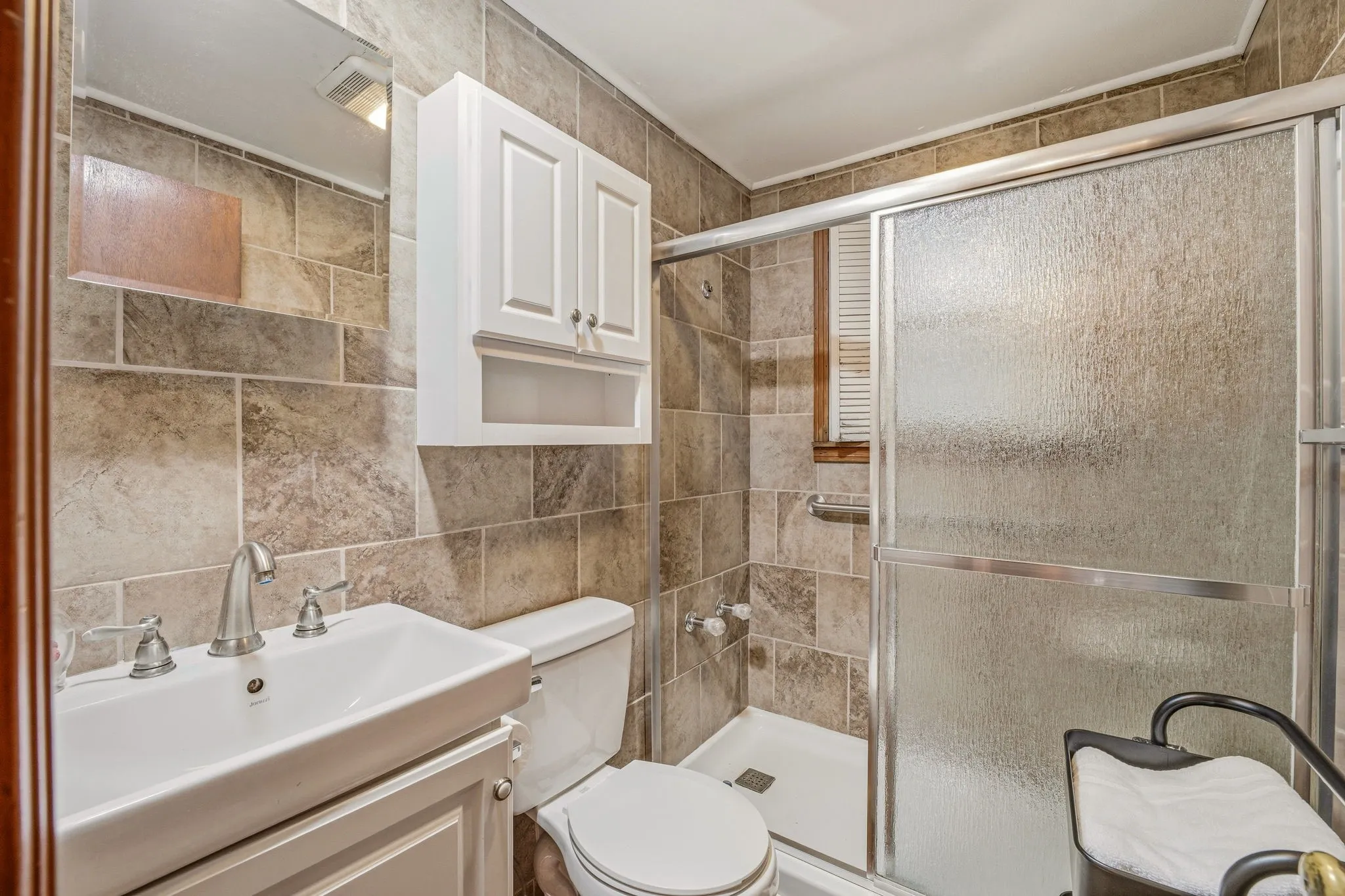 Bathroom featuring full wall tiling, a white sink vanity with brushed nickel fixtures, a white cabinet with shelving, and a glass-enclosed shower with a white shower pan