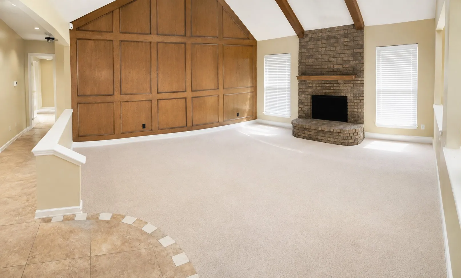 Vaulted ceiling with exposed wood beams, paneled wood accent wall, brick fireplace with mantel, two windows with blinds, and carpeted flooring. These pictures are prior to the current tenant moving in.