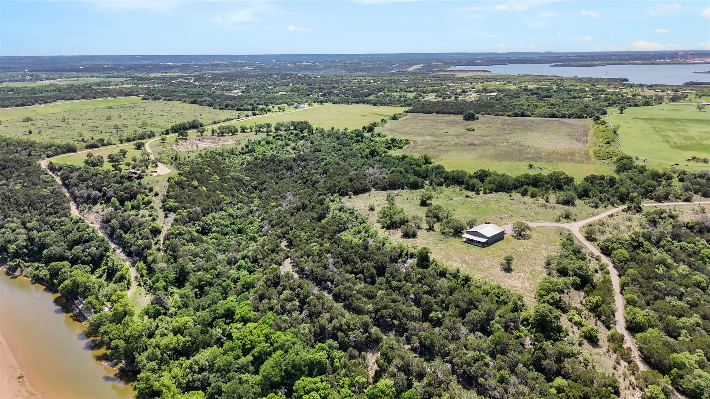 Expansive rural landscape featuring a body of water, extensive tree coverage, open fields, and a distant building
