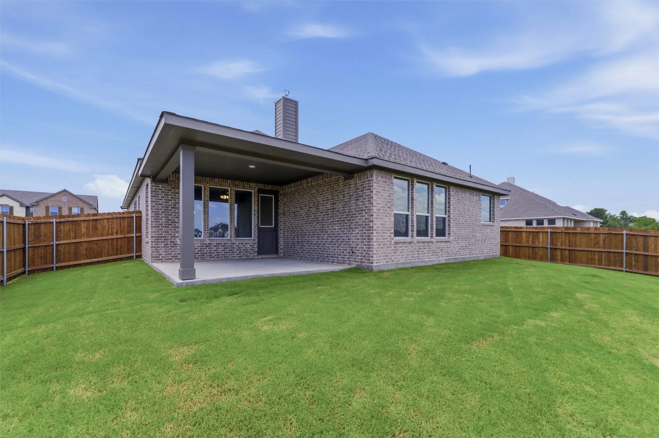 Expansive green lawn surrounds a brick exterior home featuring a covered concrete patio, wood privacy fencing, and multiple windows