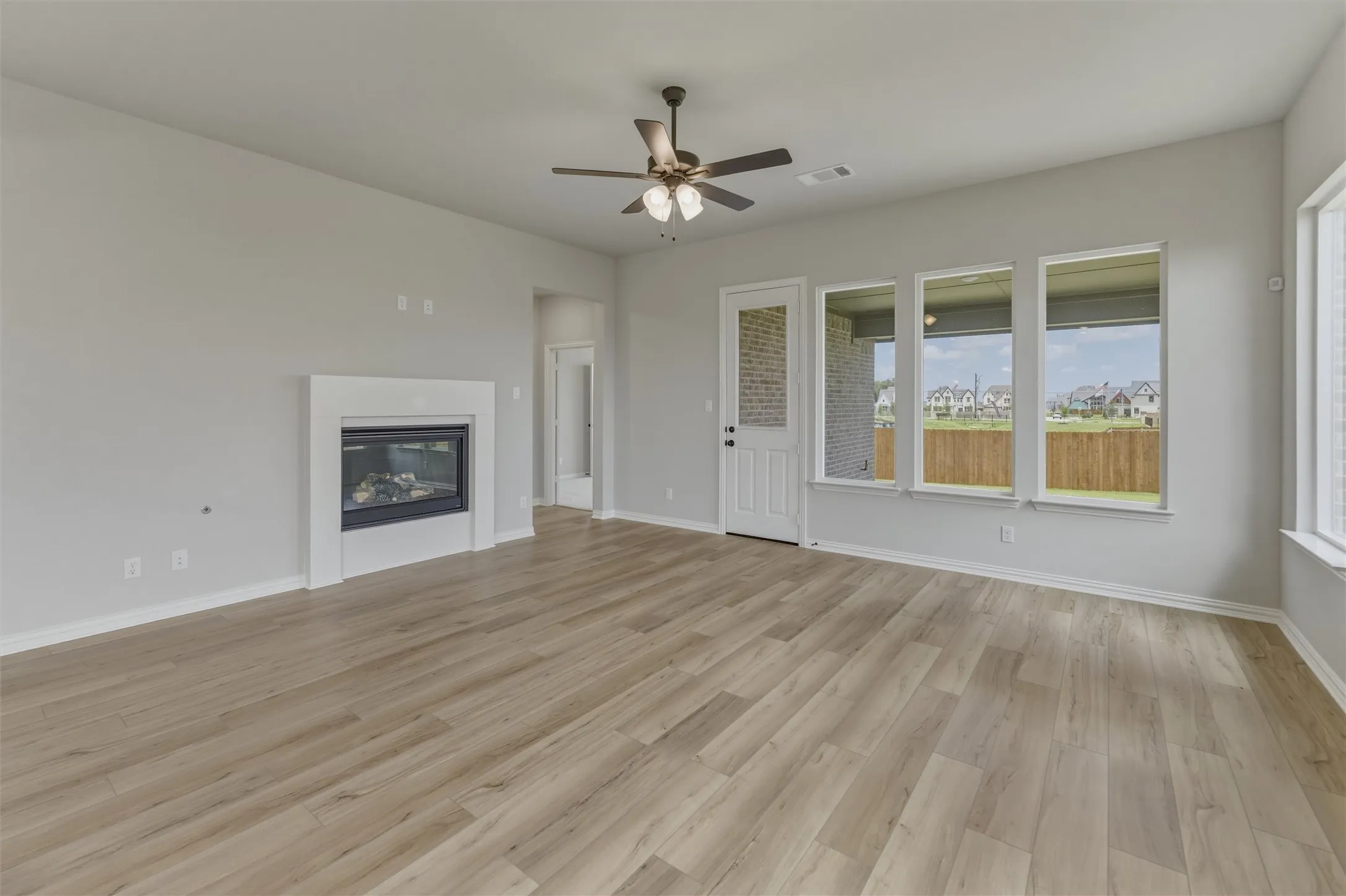 Spacious interior featuring wood-finish flooring, a contemporary fireplace with a white mantel, multiple large windows, and a ceiling fan with integrated lighting