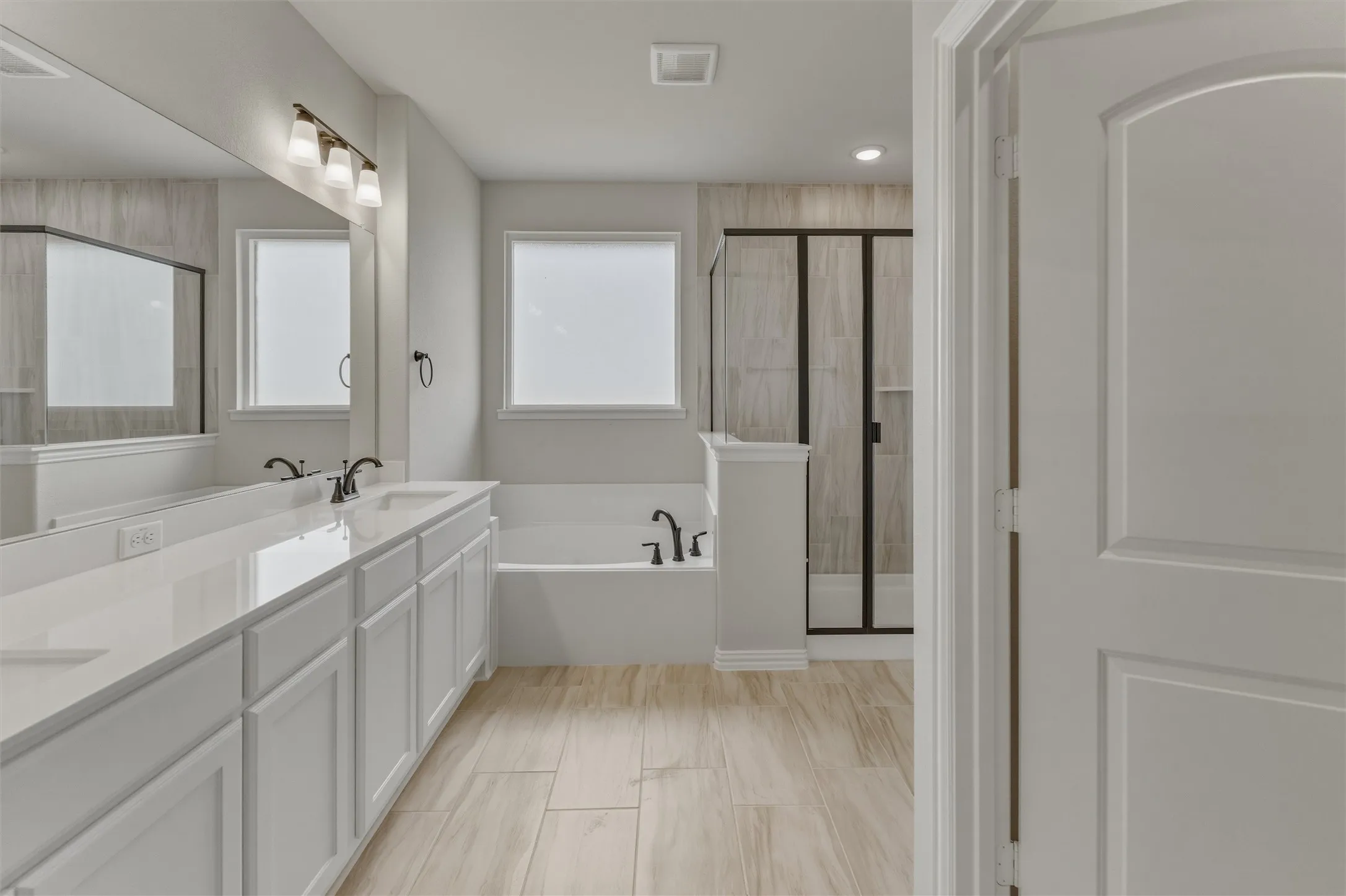 Spacious bathroom featuring a dual vanity with white cabinetry and light-toned countertops, a built-in tub, a separate shower with dark-framed glass enclosure, and light wood-look tile flooring