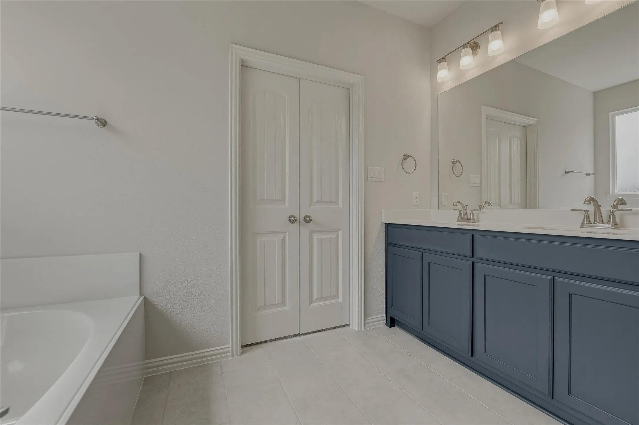 Dual-sink vanity with blue cabinetry and white countertops, large wall-mounted mirror, and a built-in bathtub