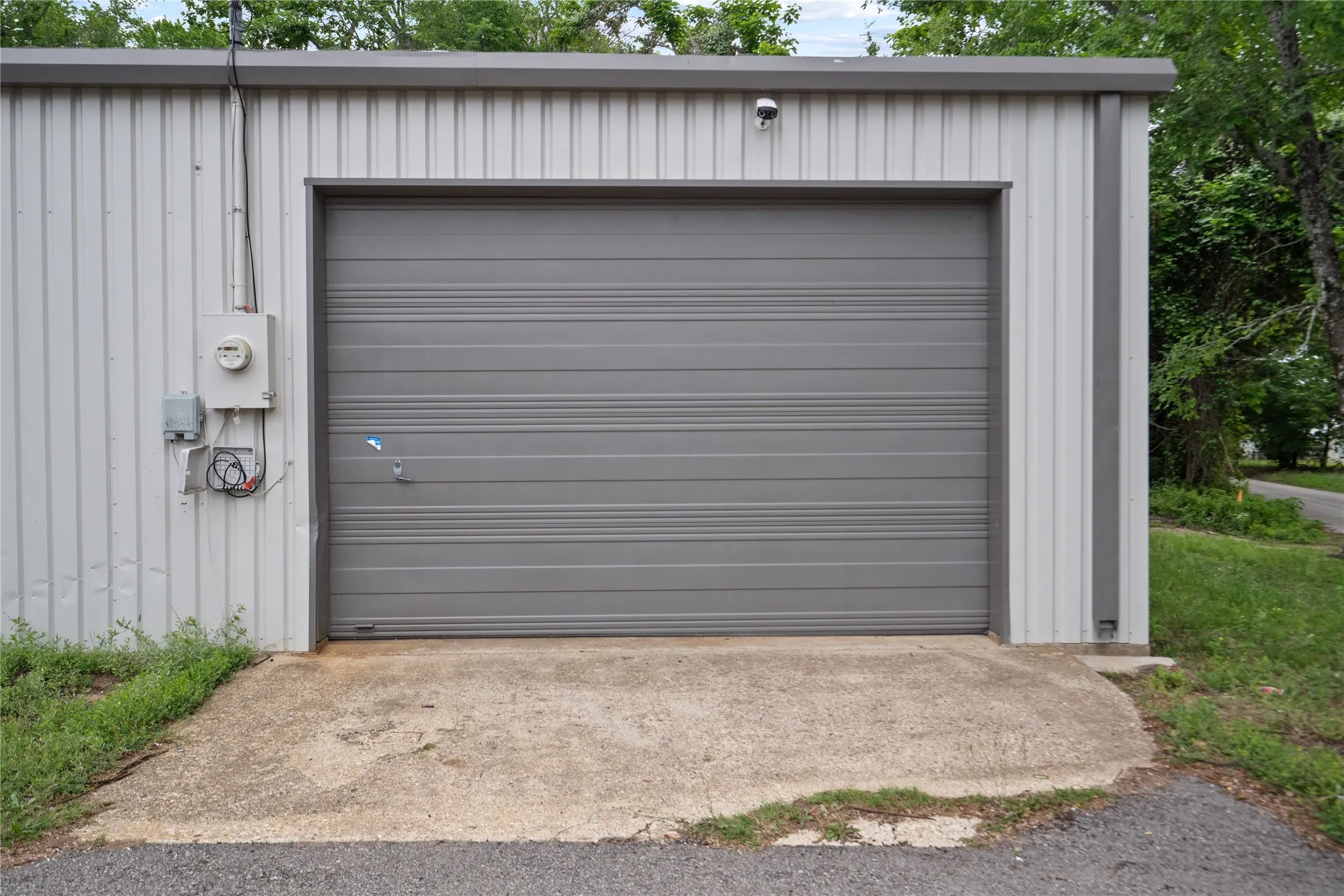 Metal-sided building featuring a gray roll-up door, exterior electrical meter, and concrete approach pad
