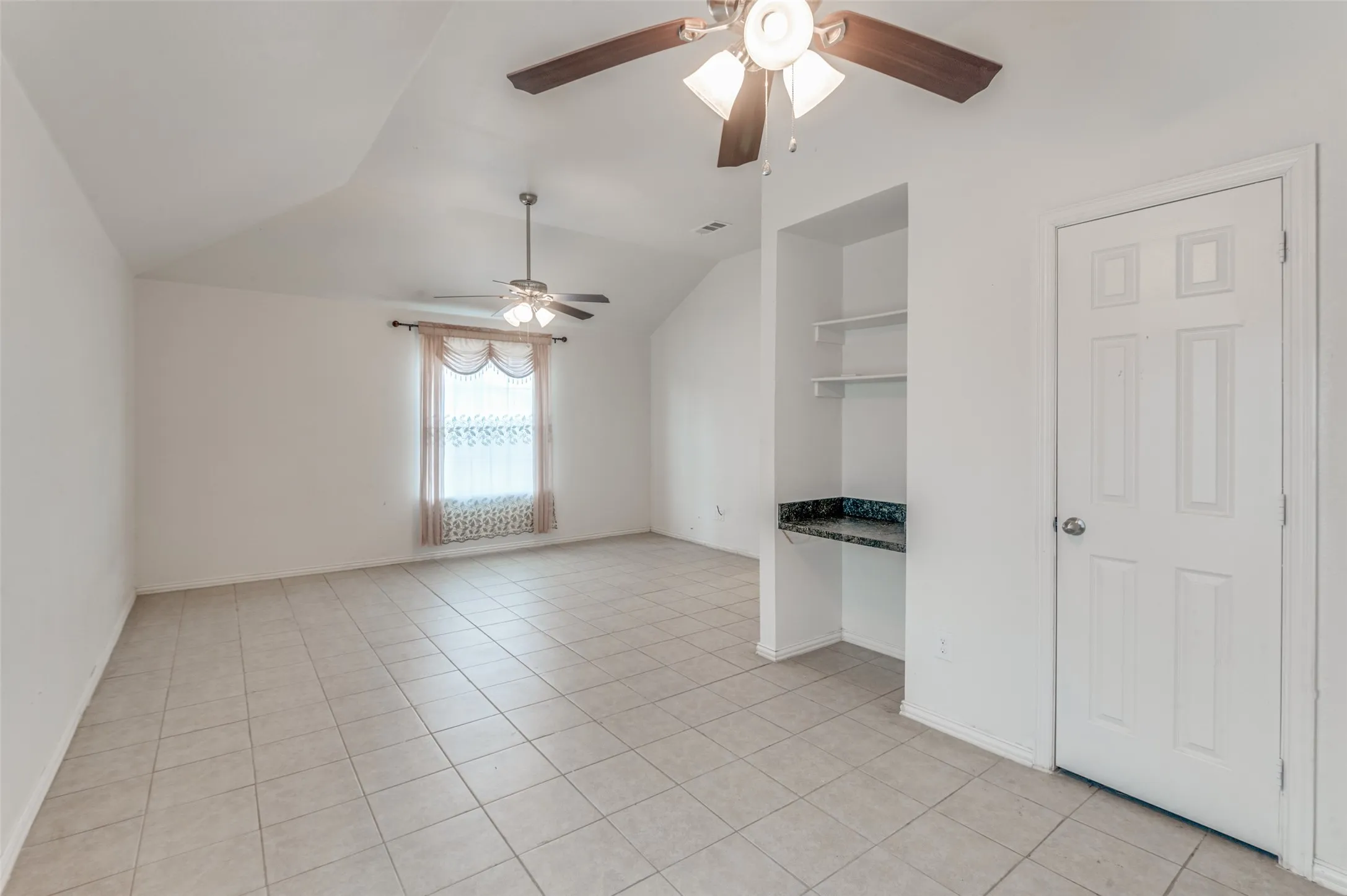 Spacious room featuring neutral tile flooring, a built-in desk with a dark patterned countertop and shelving, a six-panel door, a window with sheer and fabric treatments, and two ceiling fans with integrated lighting