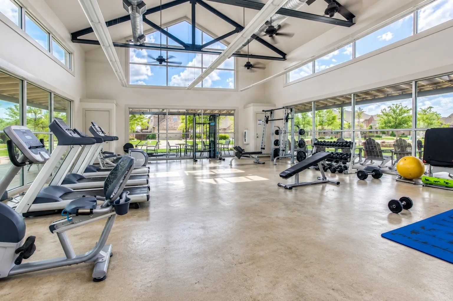 Spacious fitness center featuring high vaulted ceilings with exposed beams, expansive windows, polished concrete flooring, and a variety of exercise equipment