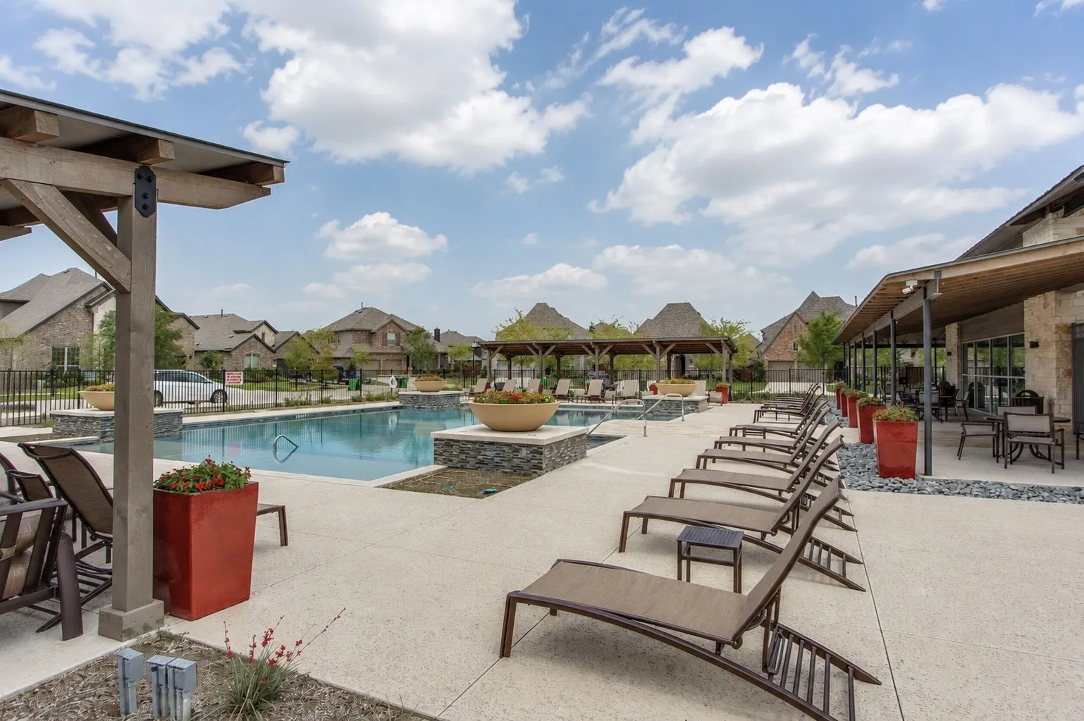Community pool area featuring a perimeter of lounge chairs, multiple shaded pergolas, and a stone-clad building with an extended covered patio