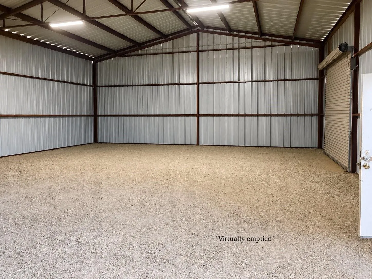 Galvanized metal wall panels, exposed structural beams, high-mounted fluorescent lighting, gravel flooring, and an overhead roll-up door