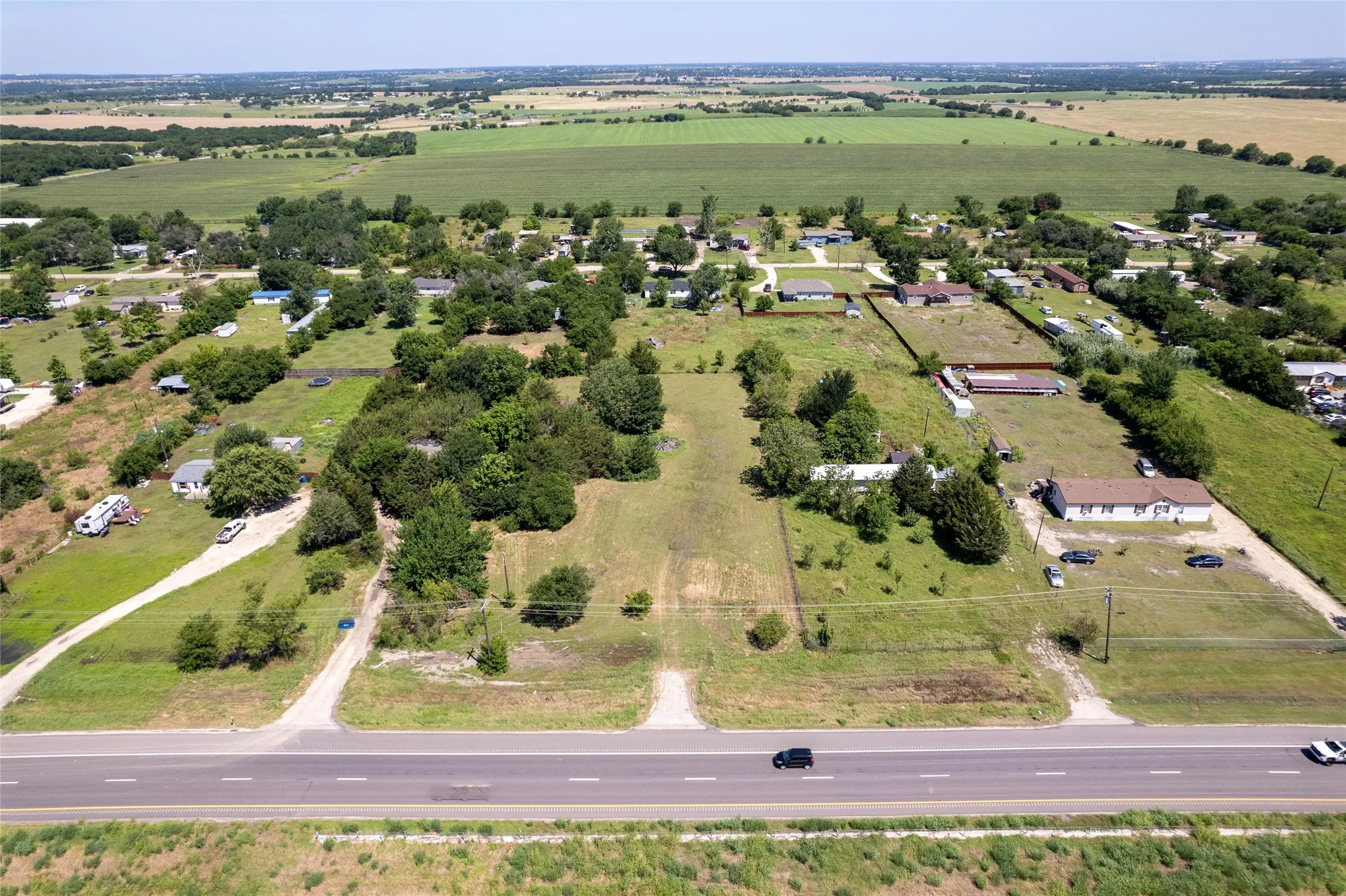 Expansive rural landscape featuring a paved roadway, multiple grass-covered parcels, mature trees, and distant cultivated fields