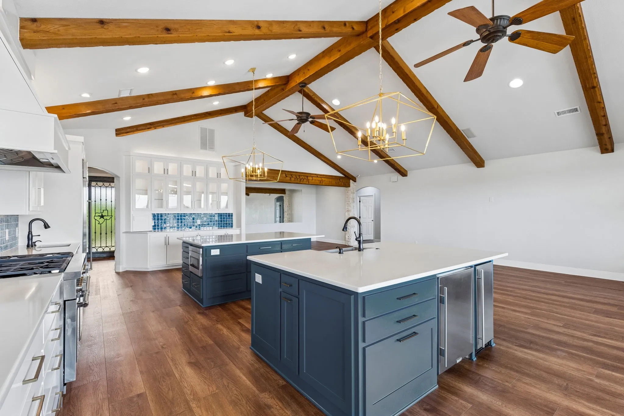 The kitchen opens under dramatic 14 ft vaulted ceilings with exposed beams, featuring two large islands designed for gathering and everyday living. A custom built-in hutch with glass cabinetry and a striking blue tile backsplash adds charm, storage, and a distinctive design moment.