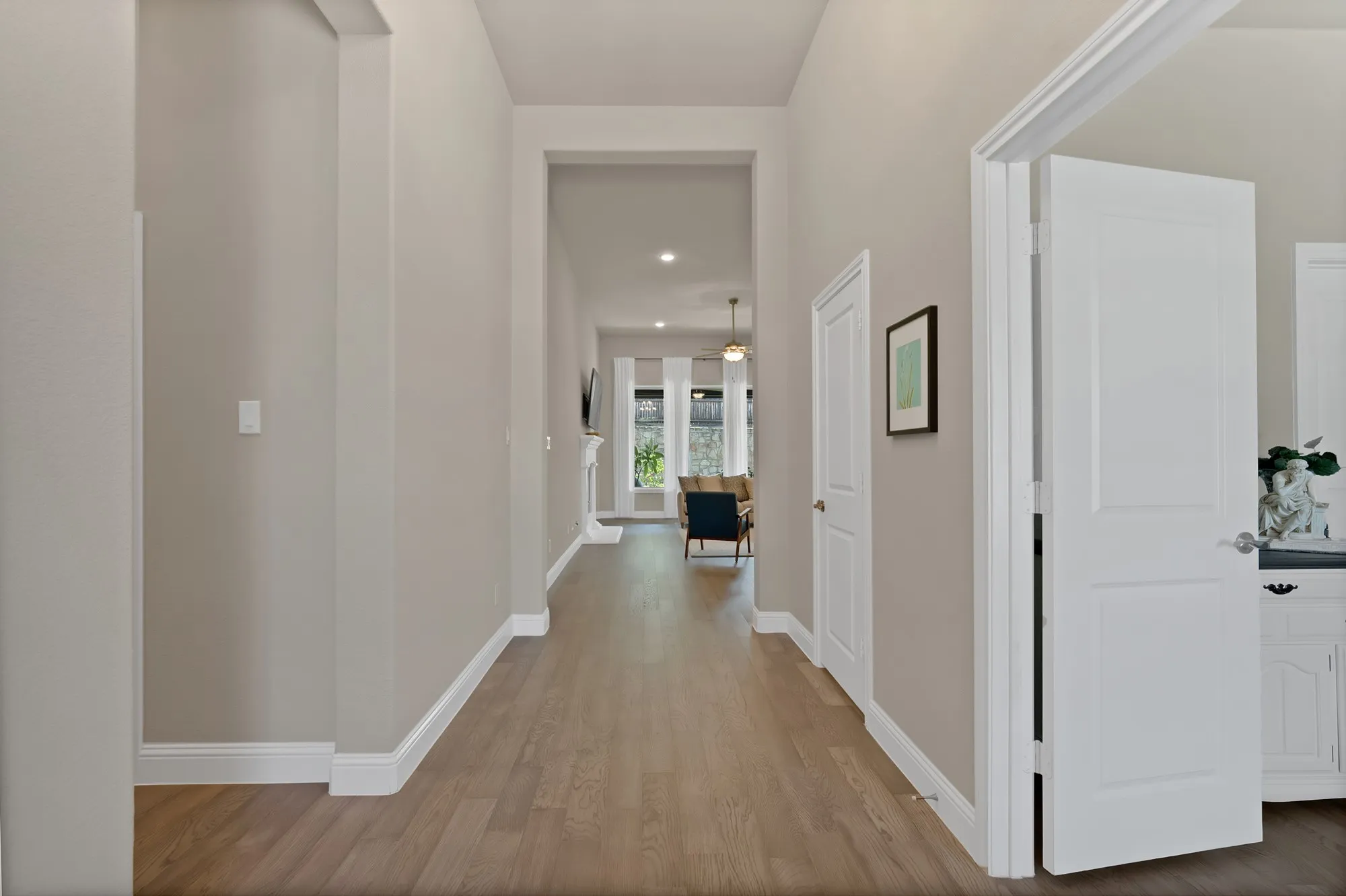 Inviting hallway with light wood-style flooring and neutral wall tones, leading to a living area with natural light from windows