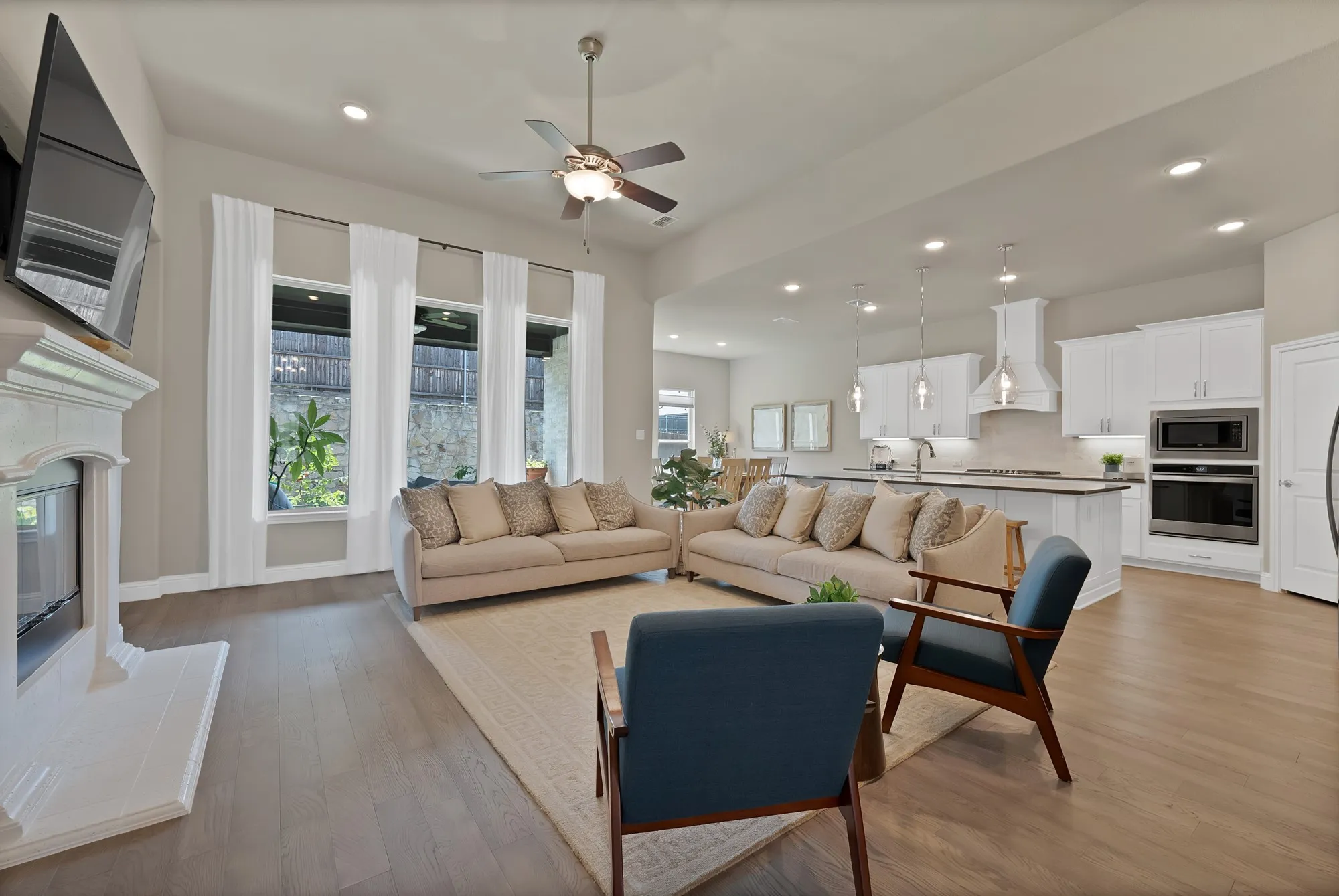 Spacious living area featuring light wood flooring, a fireplace with a white mantle, and large windows with white curtains