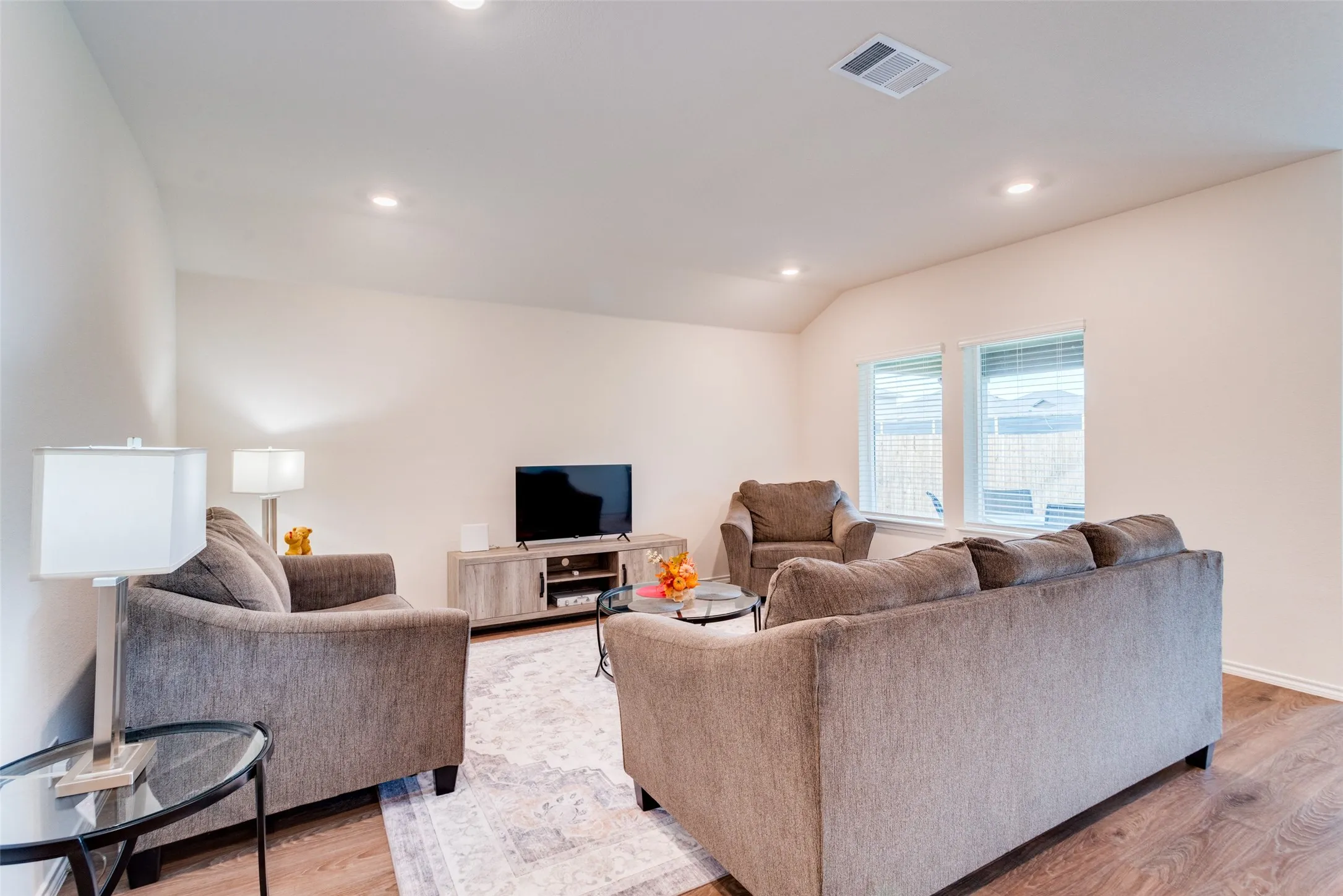 Spacious living area featuring recessed lighting, wood-finish flooring, two windows with blinds, a light-colored accent wall, and a ceiling-mounted air vent