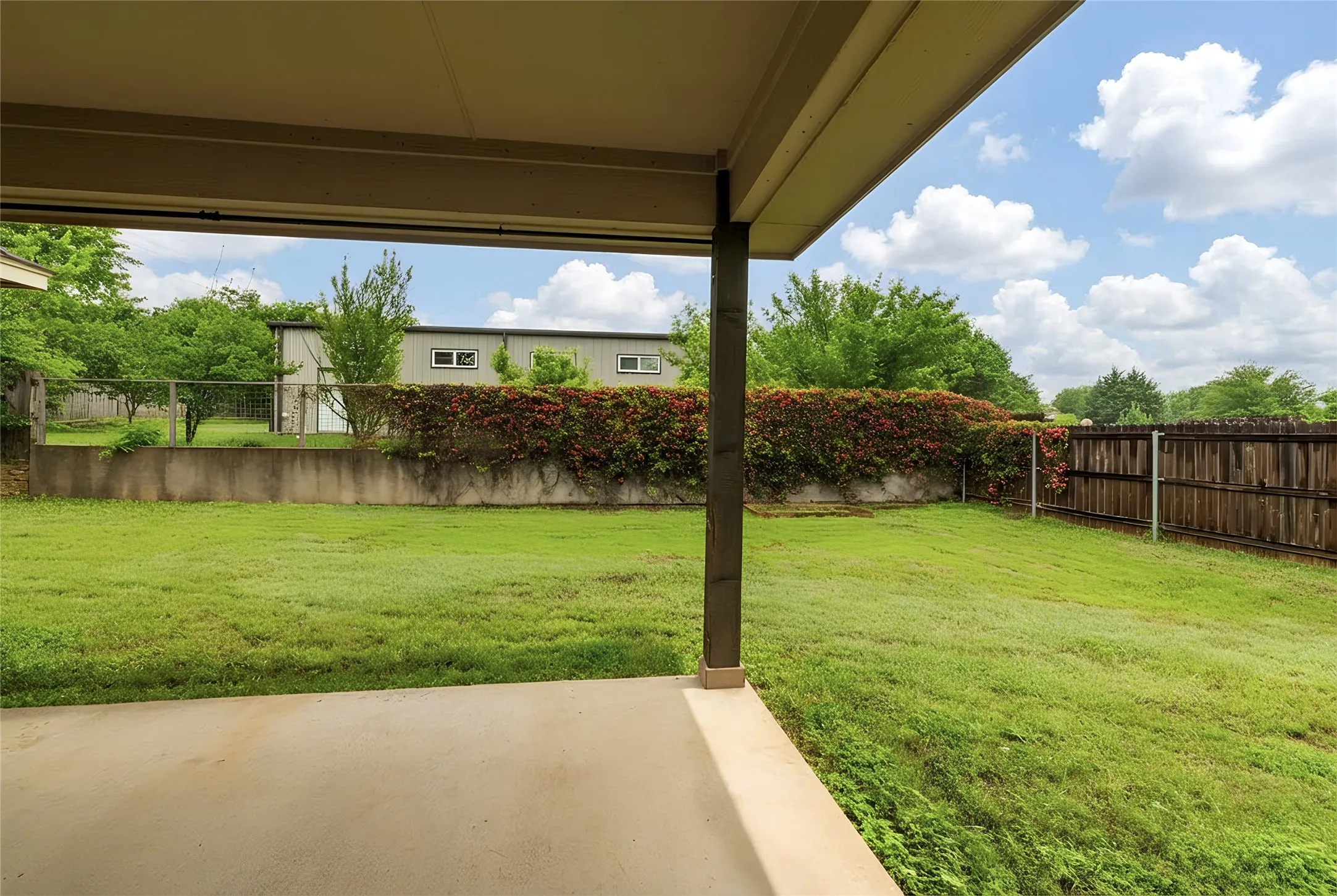 Covered patio with a concrete surface, featuring a green lawn, established perimeter foliage, concrete retaining wall, and a wood fence
