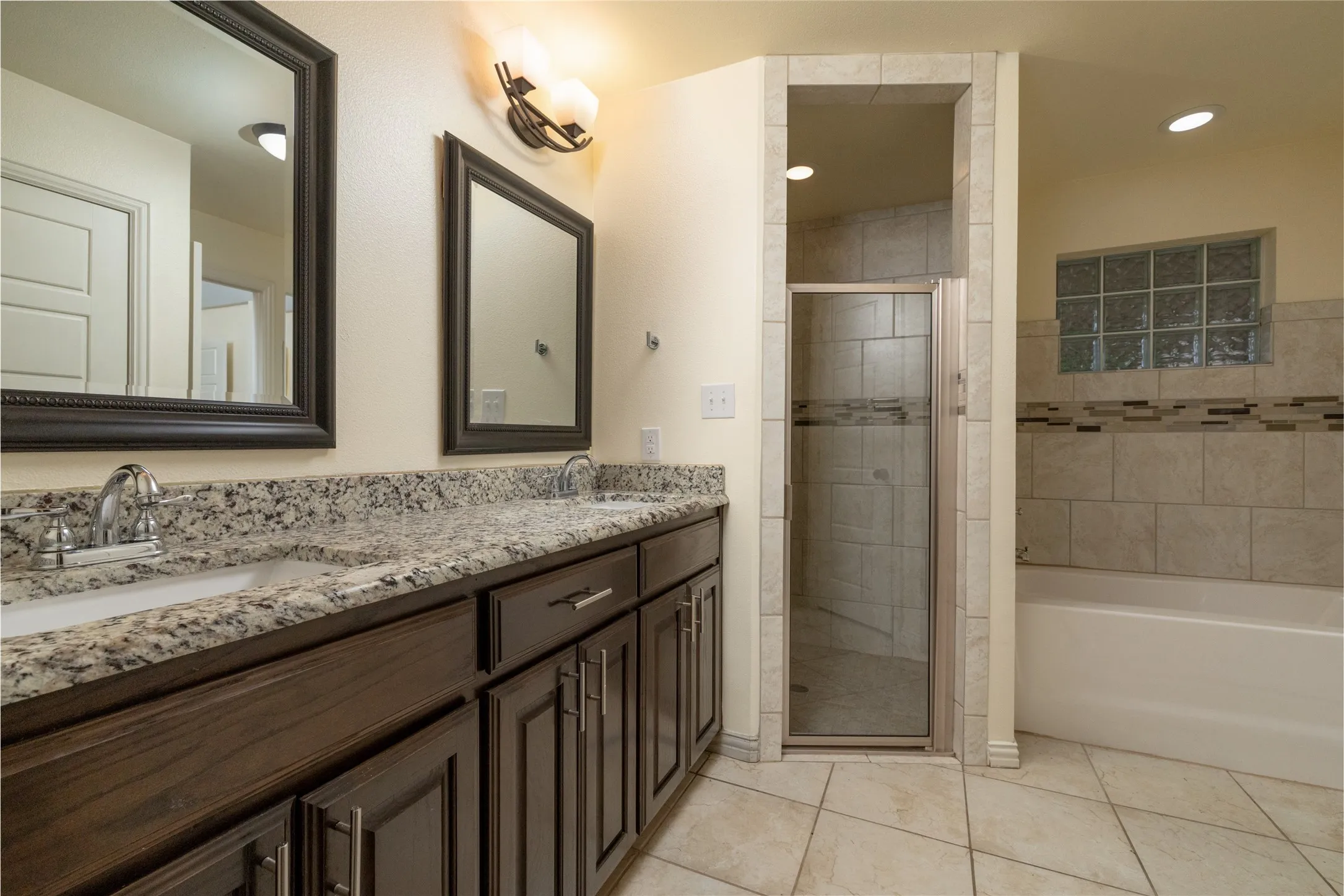 Bathroom featuring a dual vanity with granite-finish countertop, dark wood cabinetry, two framed mirrors, a glass-enclosed shower, and a separate bathtub with tile surround and glass block window