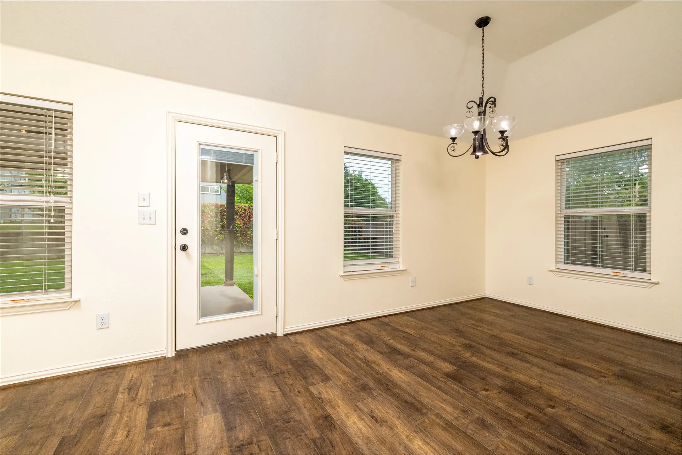 Spacious room featuring vaulted ceilings, wood-finish flooring, a glass-paneled exterior door, and multiple windows with blinds