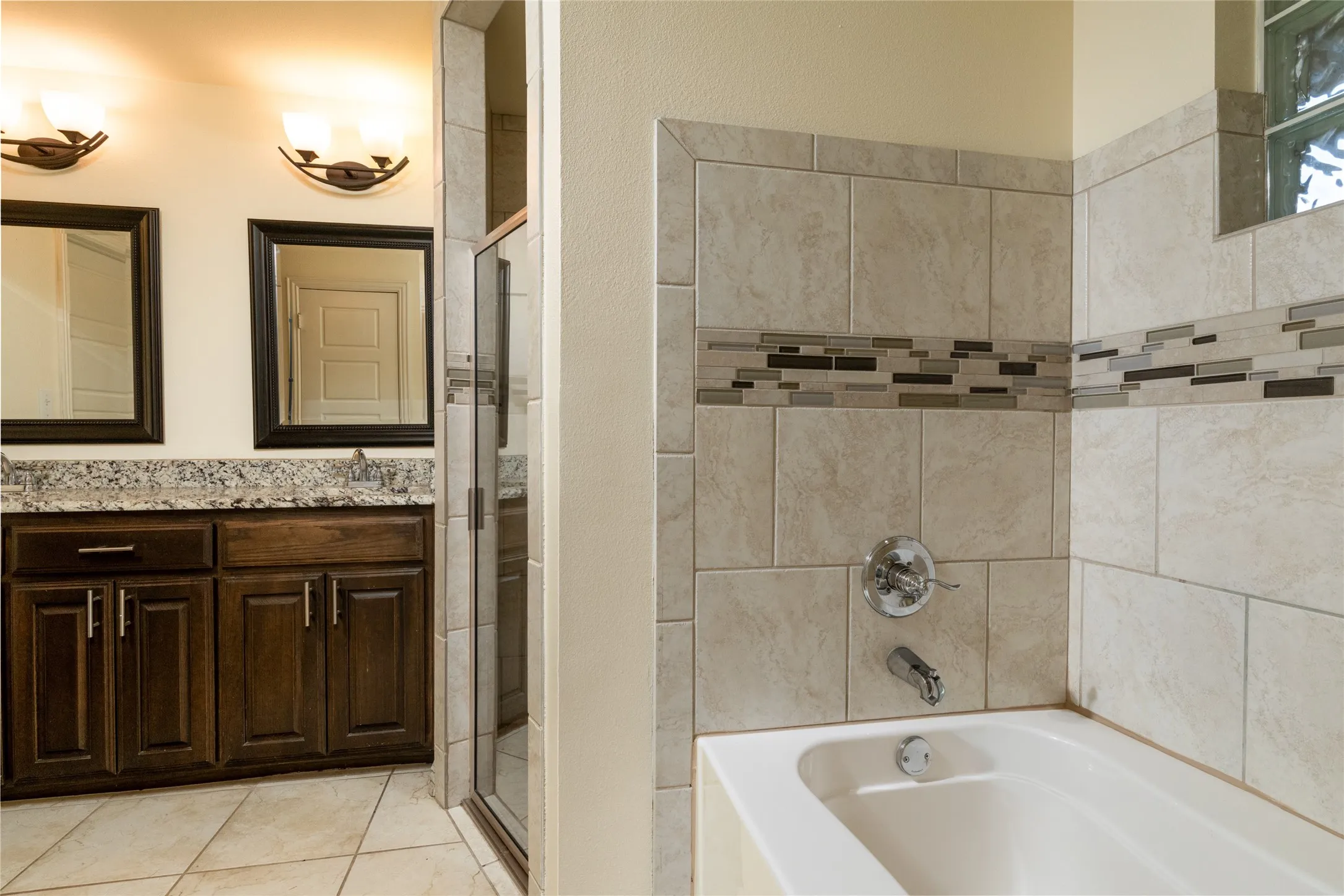 Bathroom featuring a tiled tub and shower surround with an accent mosaic border, a glass block window, a dual vanity with granite countertops, and dark wood cabinetry