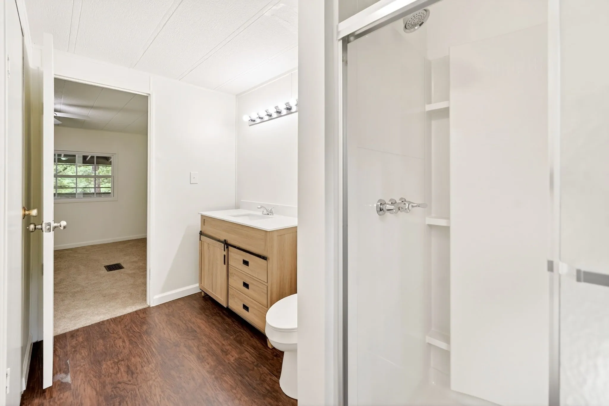 Bathroom featuring wood-finish flooring, a light wood vanity with a white countertop and integrated sink, a toilet, and a glass-enclosed shower with built-in shelving