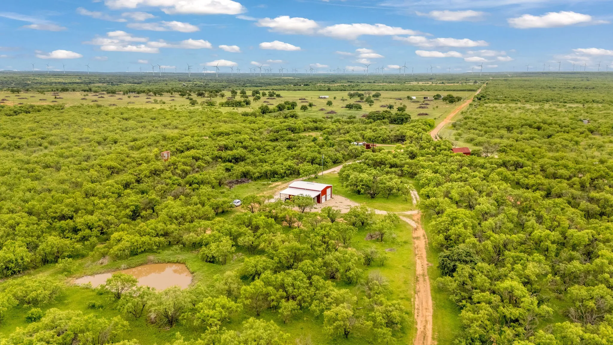 Expansive rural landscape featuring a red barn structure, a winding dirt road, a pond, abundant green foliage, and open fields extending to the horizon
